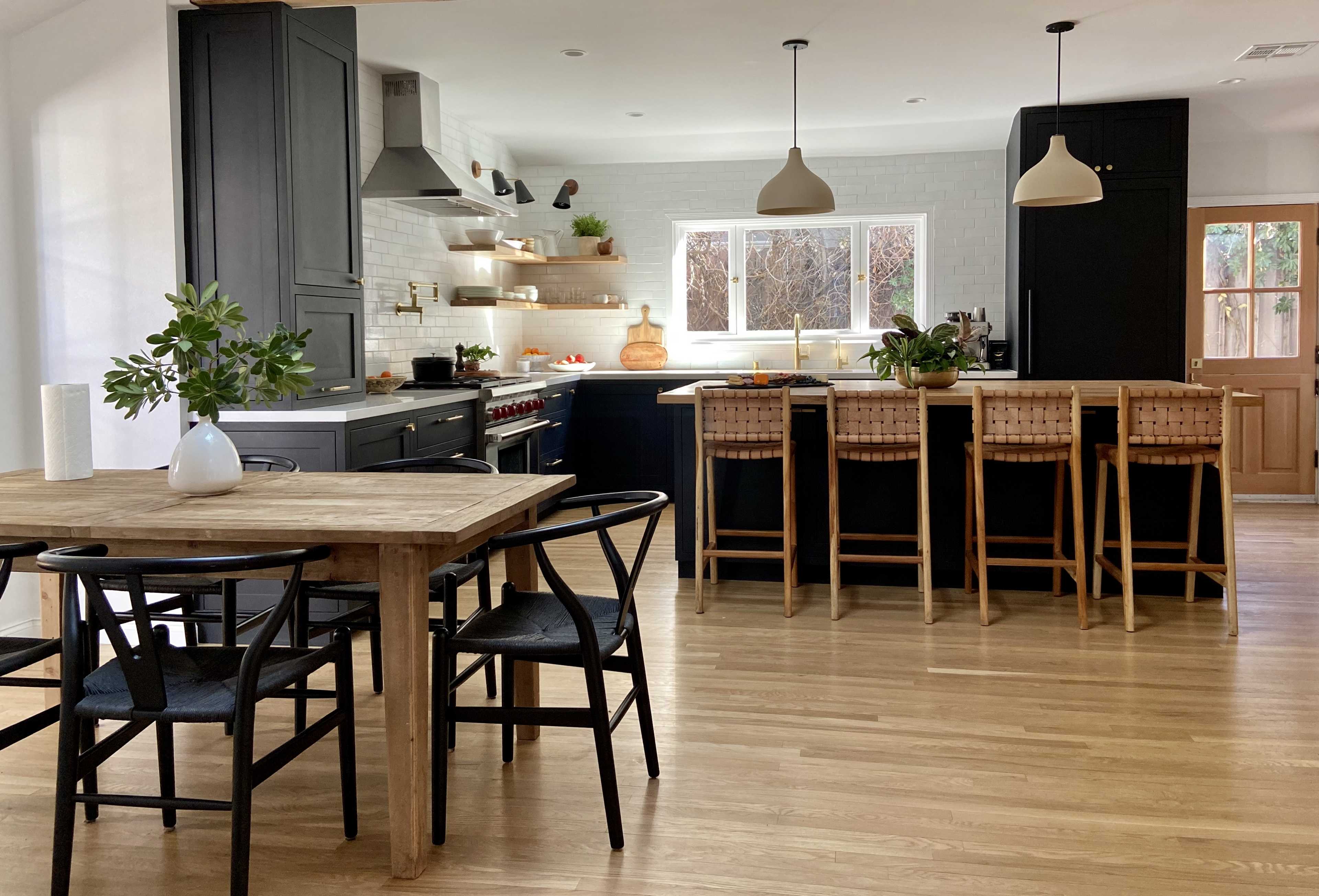 A modern kitchen and dining area with a wooden table, black chairs, a central island with bar stools, and white walls featuring shelves and a window.