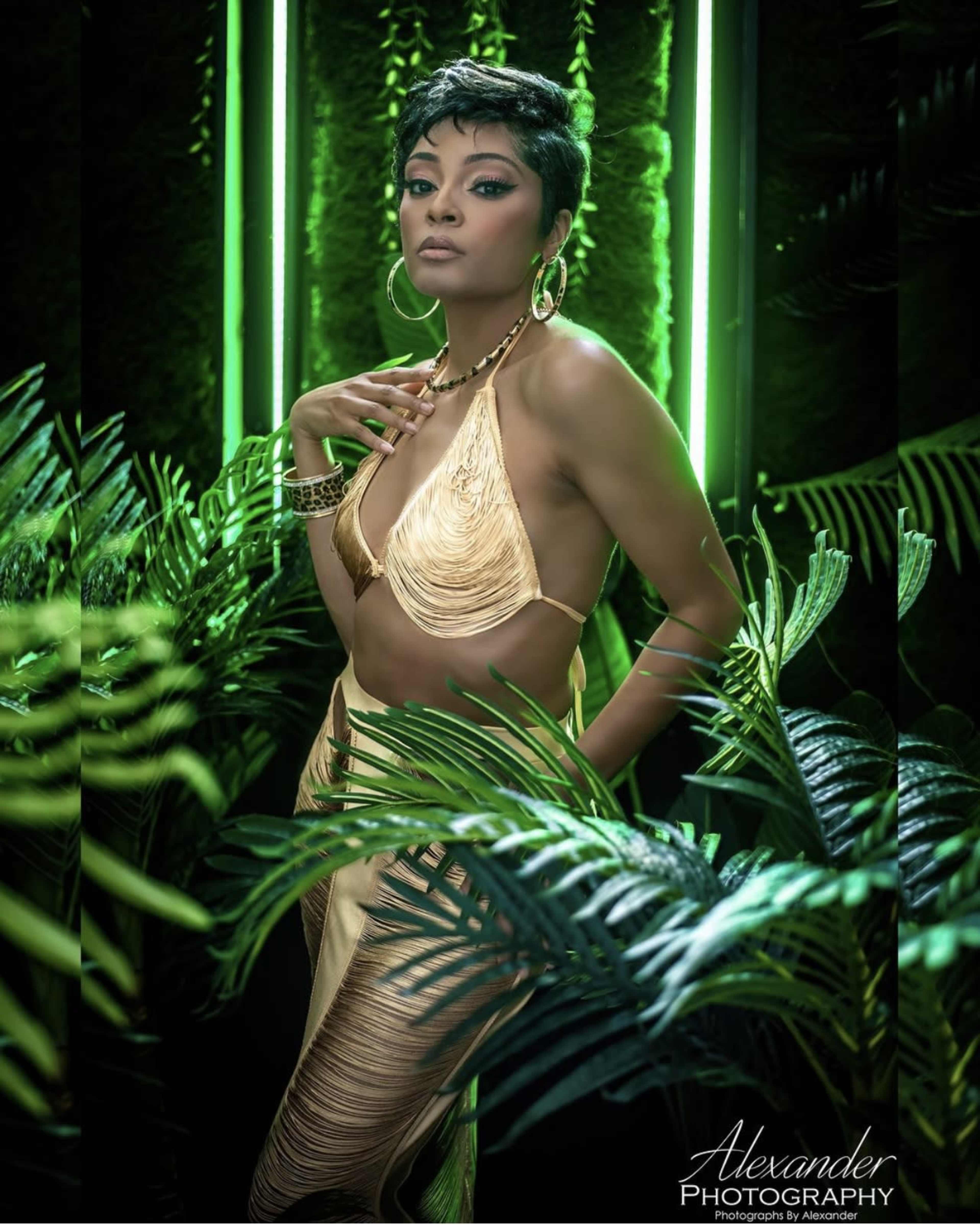 A woman in a gold outfit stands among lush green plants, illuminated by glowing neon lights.