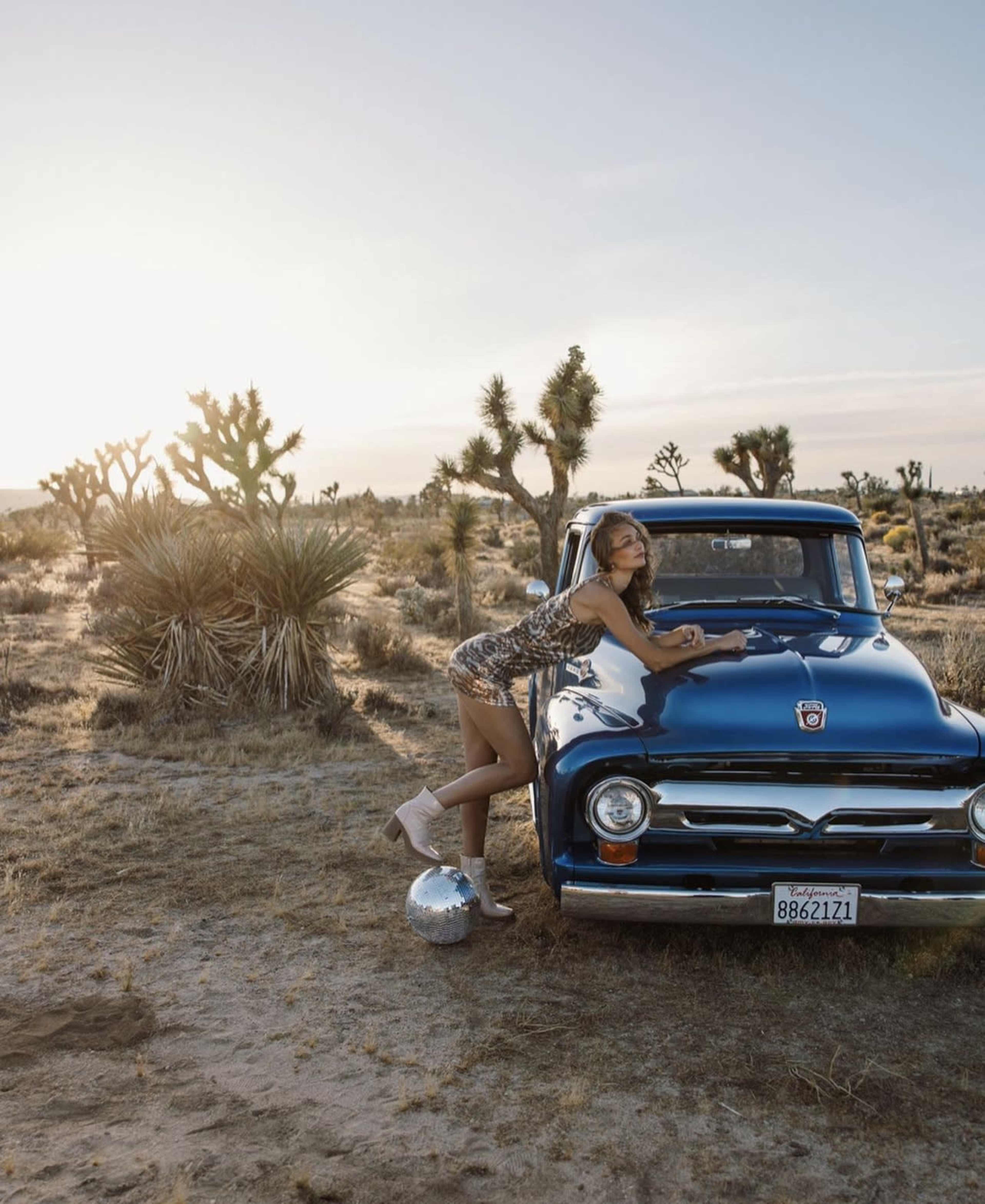 A woman in a patterned dress leans against a blue vintage truck parked in a desert landscape with Joshua trees at sunset.