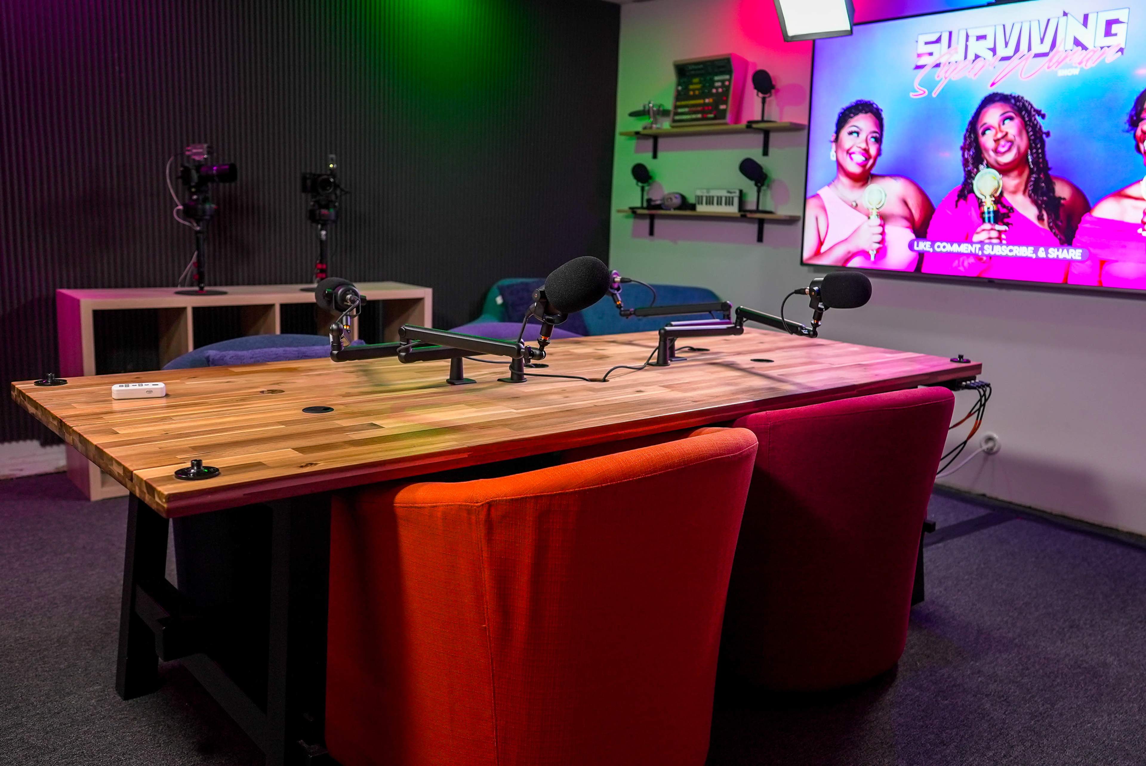 A podcast studio setup with a wooden table equipped with microphones and colorful chairs, alongside a screen displaying a show called "Surviving."