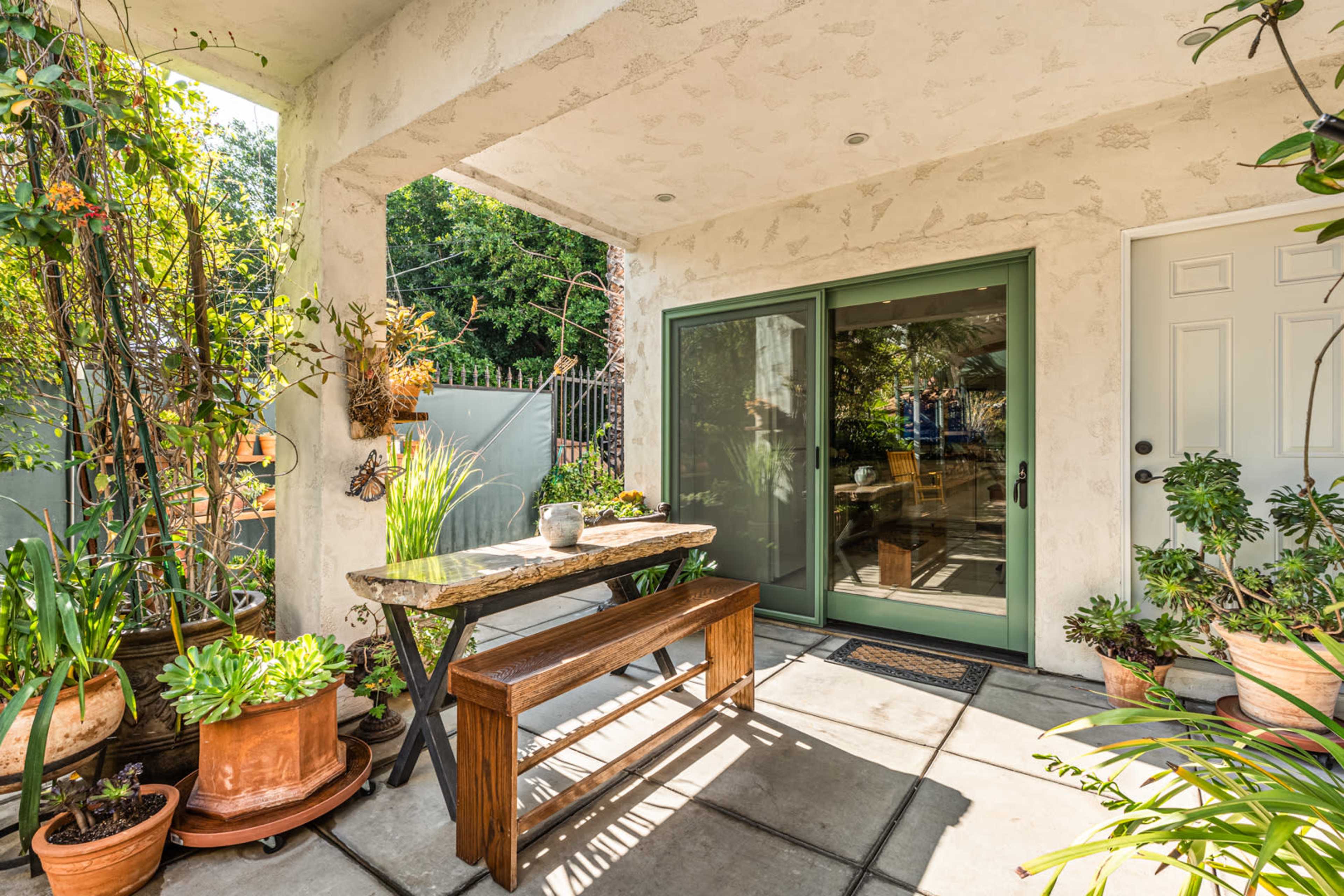 A porch area with a wooden table surrounded by various potted plants and a glass sliding door leading into a brighter room.