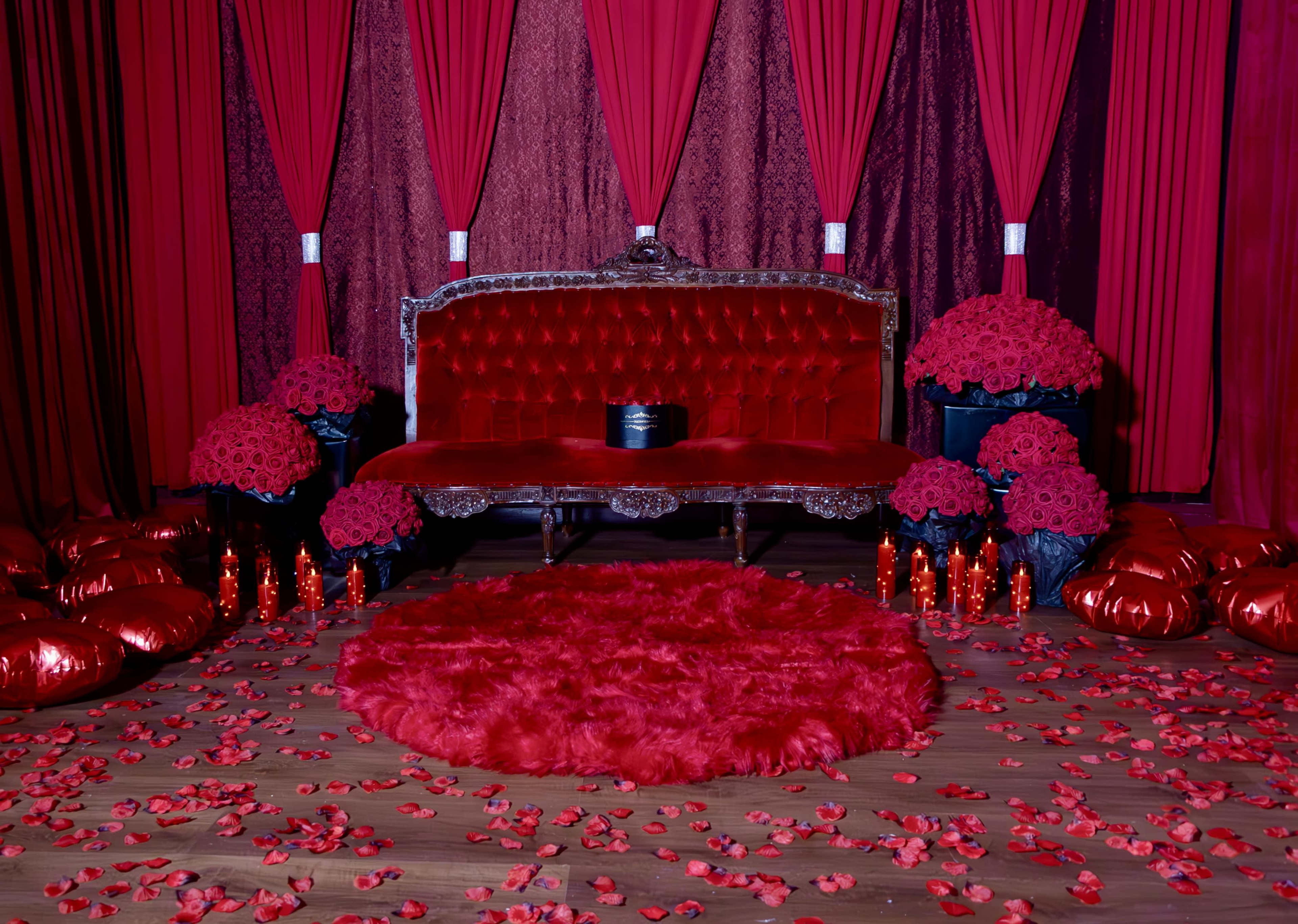 A red-themed room features a velvet sofa, large floral arrangements of roses, decorative pillows, and a circular rug surrounded by scattered rose petals and candles.