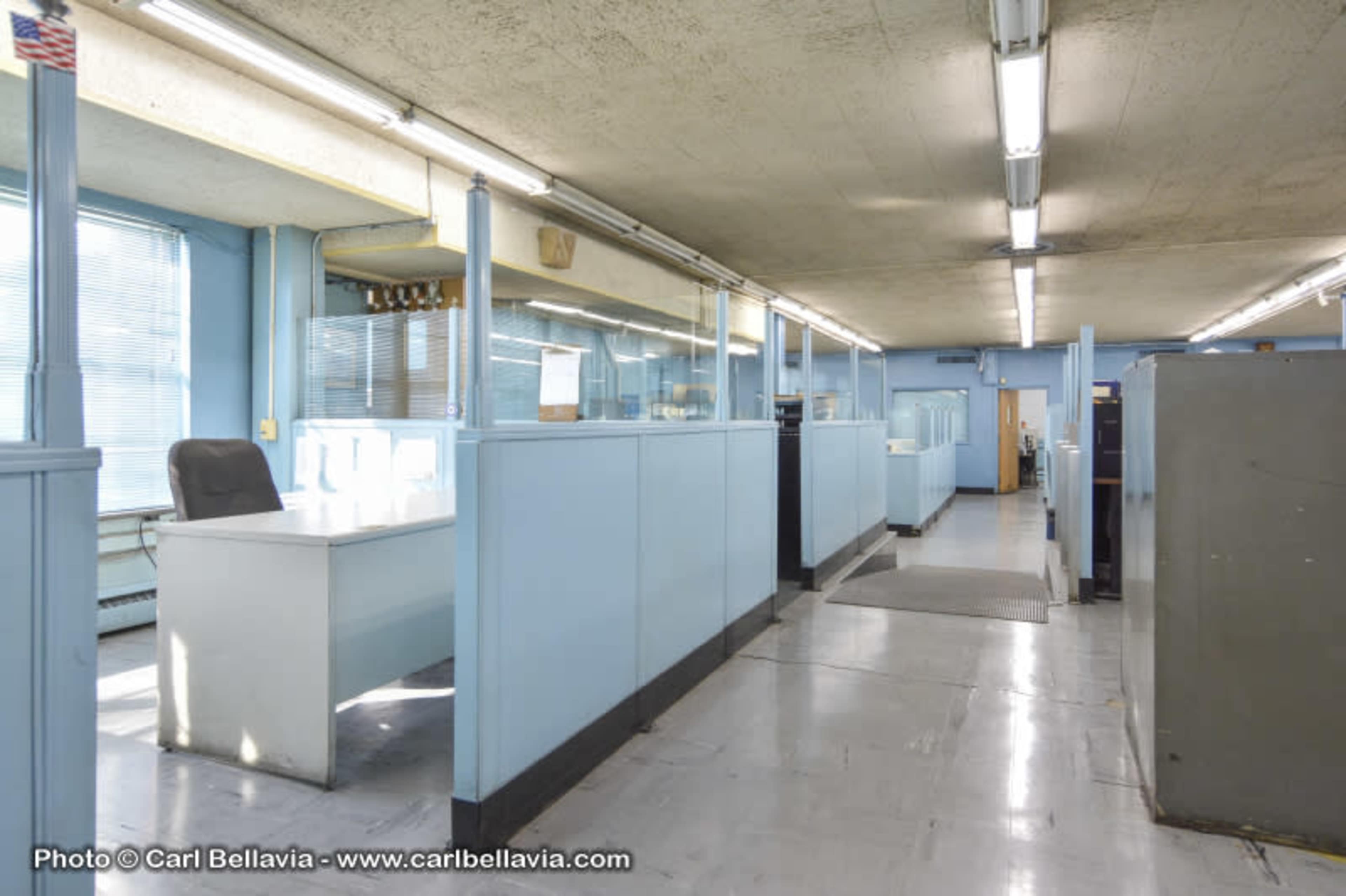 A spacious office interior with light blue walls, divided workstations, and artificial lighting.