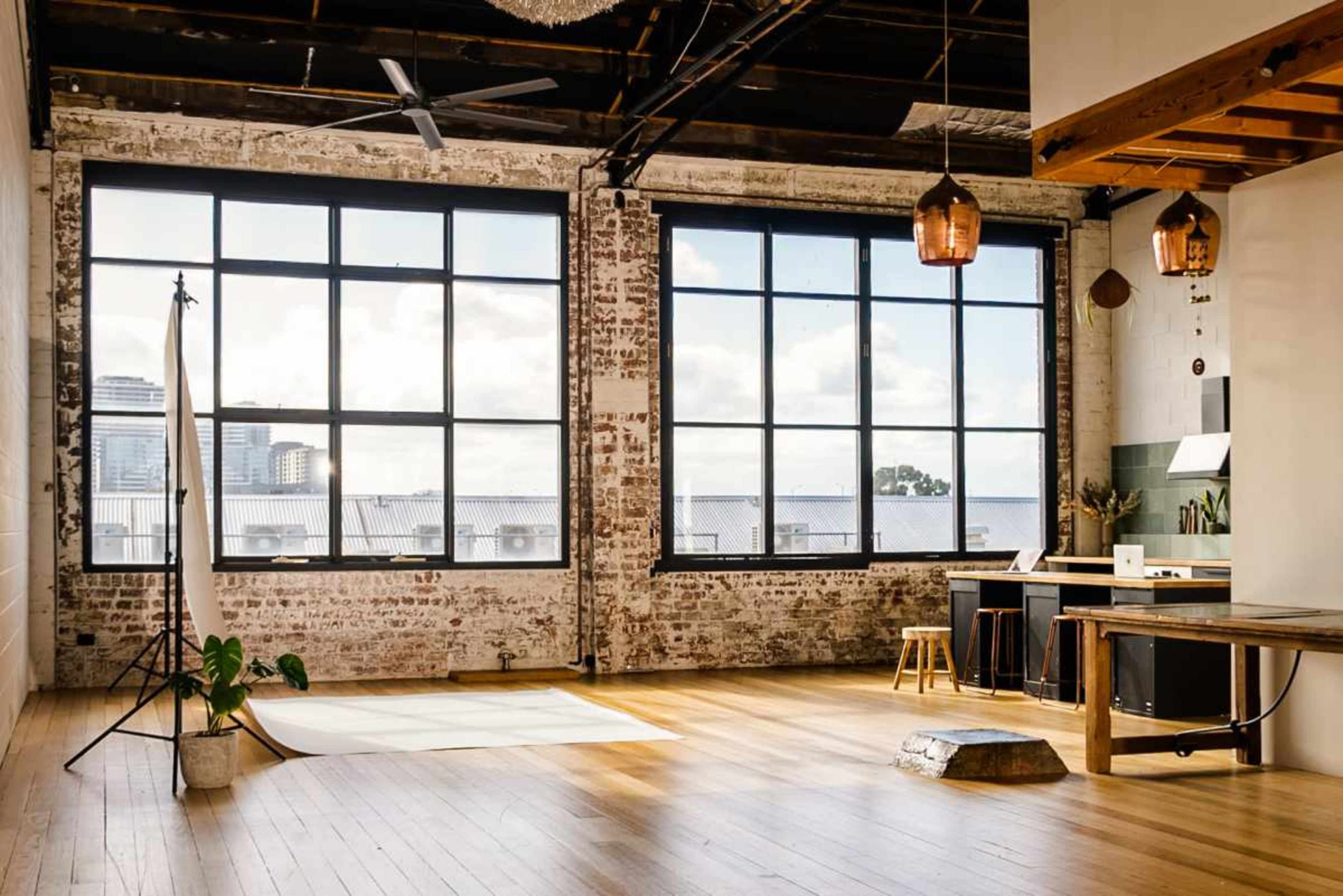 The image shows a spacious, industrial-style room with large windows, exposed brick walls, wooden flooring, a simple kitchen area, and a small green plant in a pot.