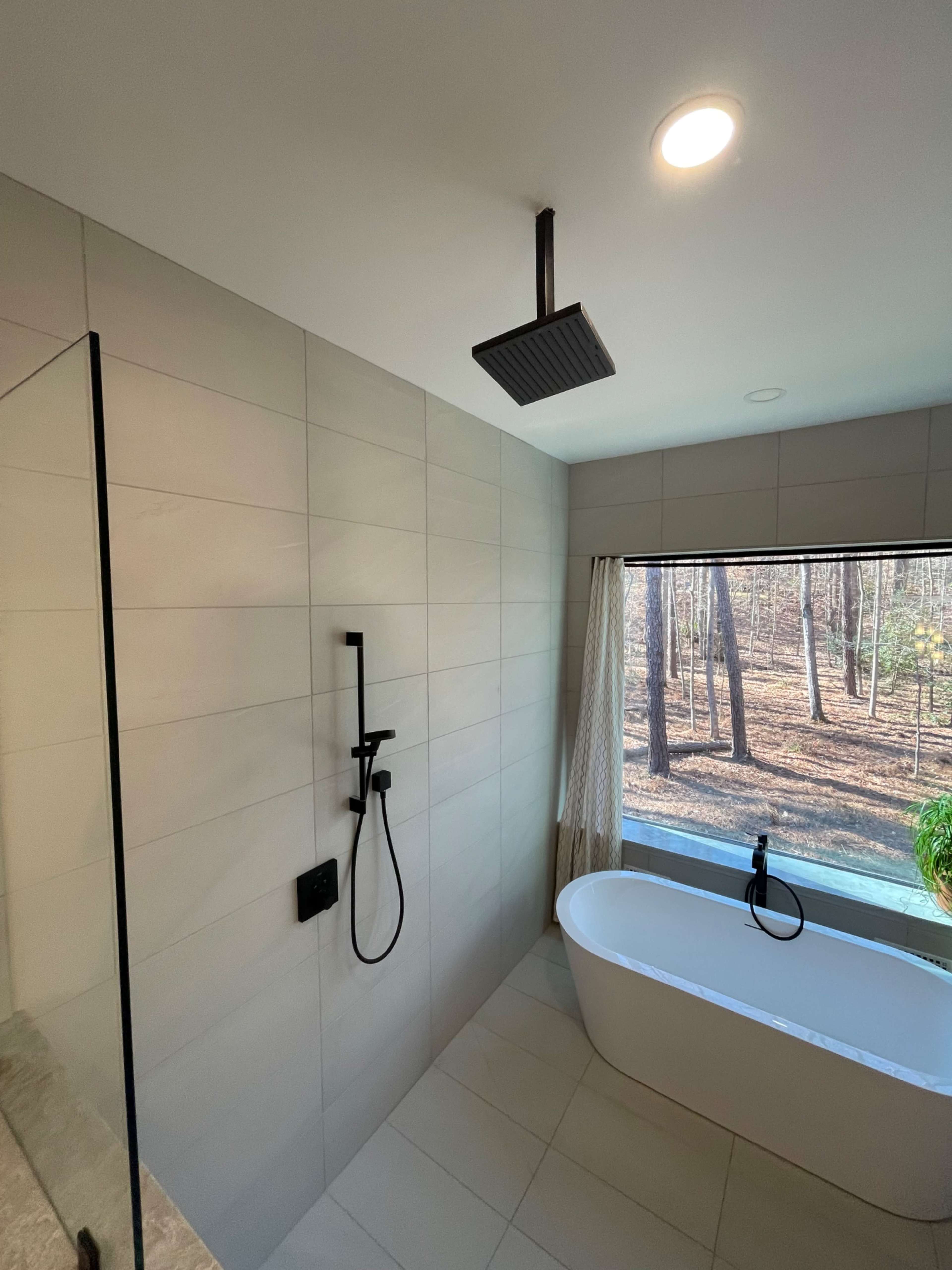 The image shows a modern bathroom featuring a freestanding tub and a walk-in shower, with a large window overlooking a wooded area.