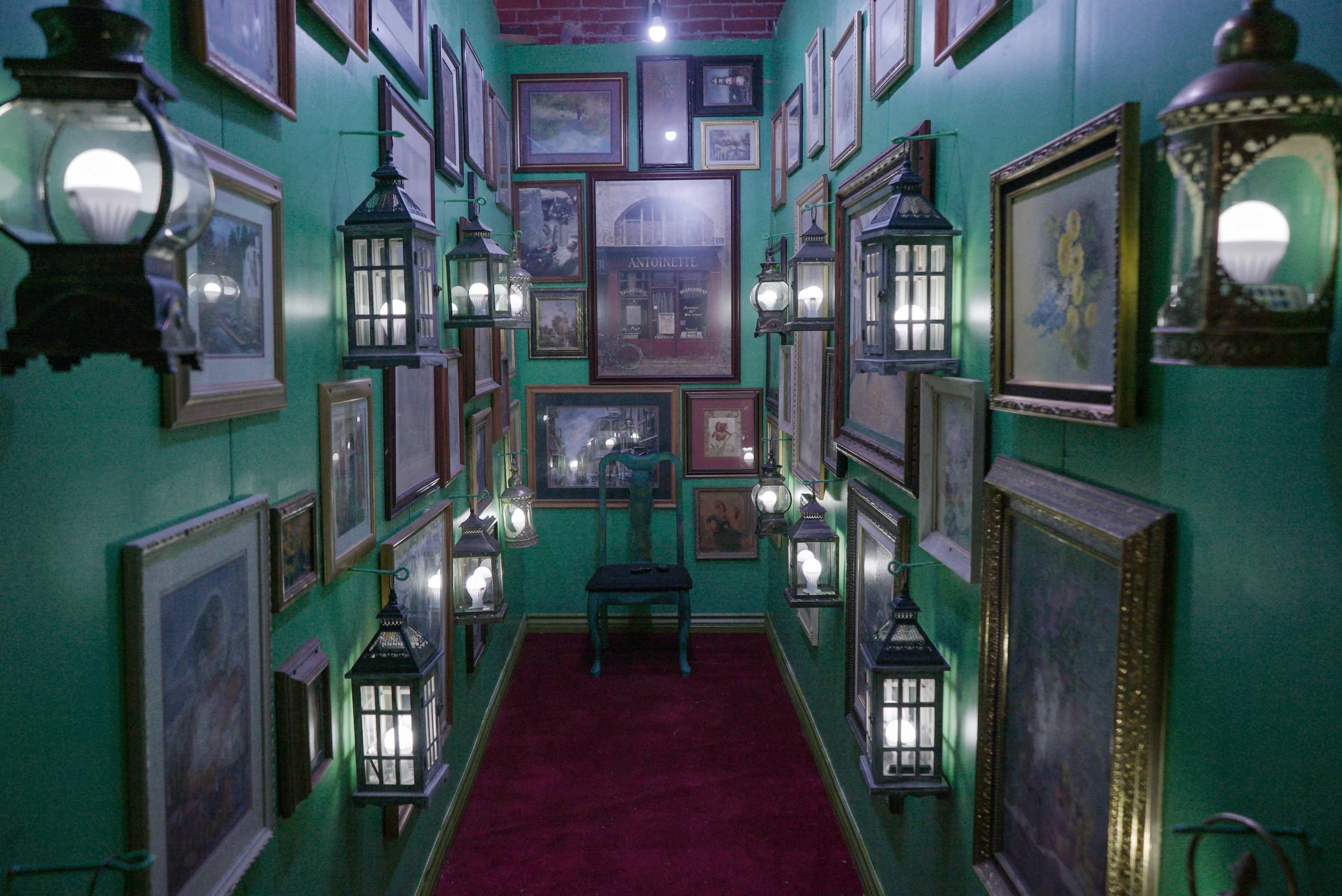 A narrow hallway lined with framed artwork and lanterns hanging from the walls, with a single chair positioned in the center.