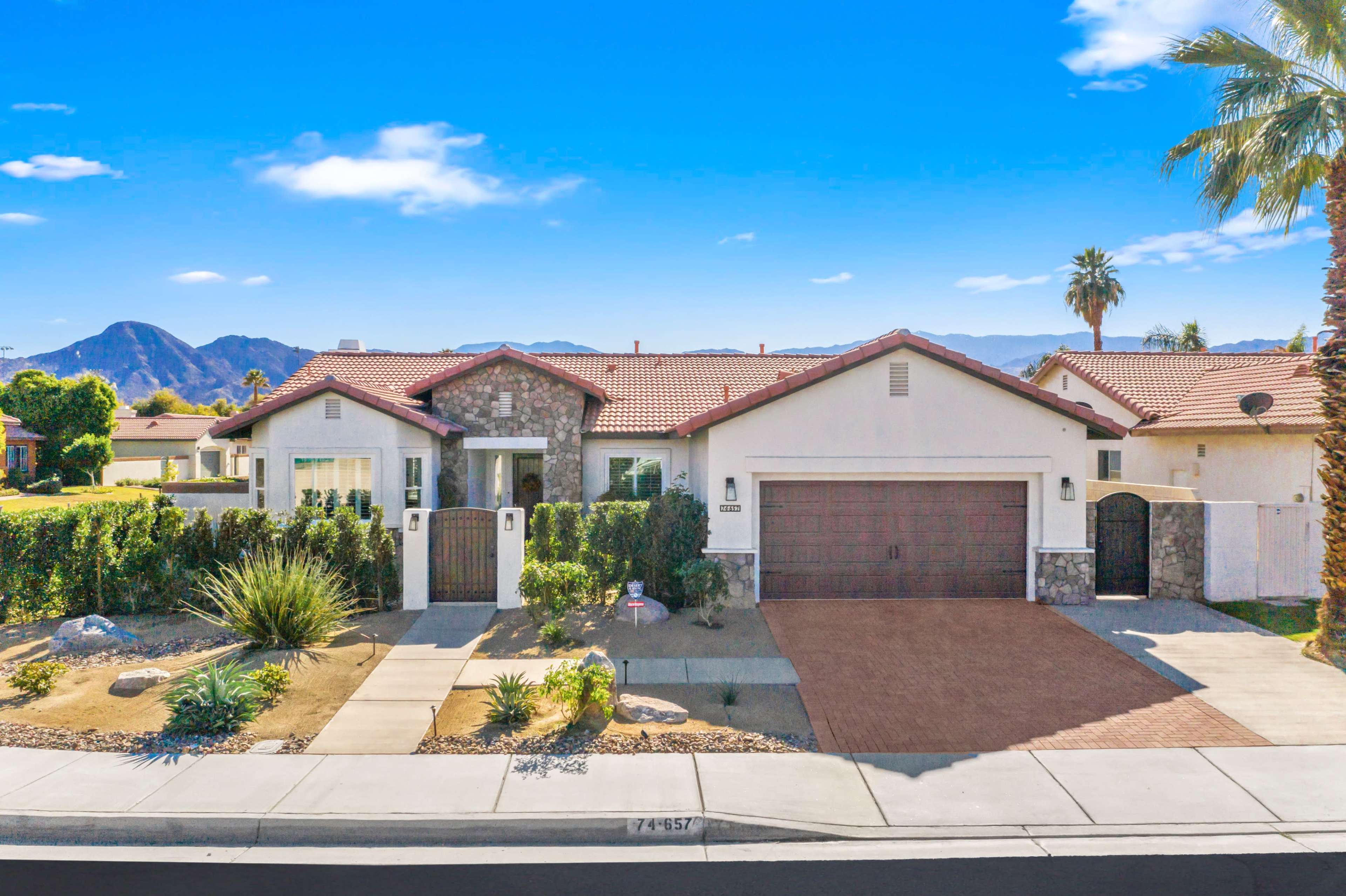 A single-story residential home with a stone-accented façade, a two-car garage, and landscaped yard, set against a backdrop of mountains.