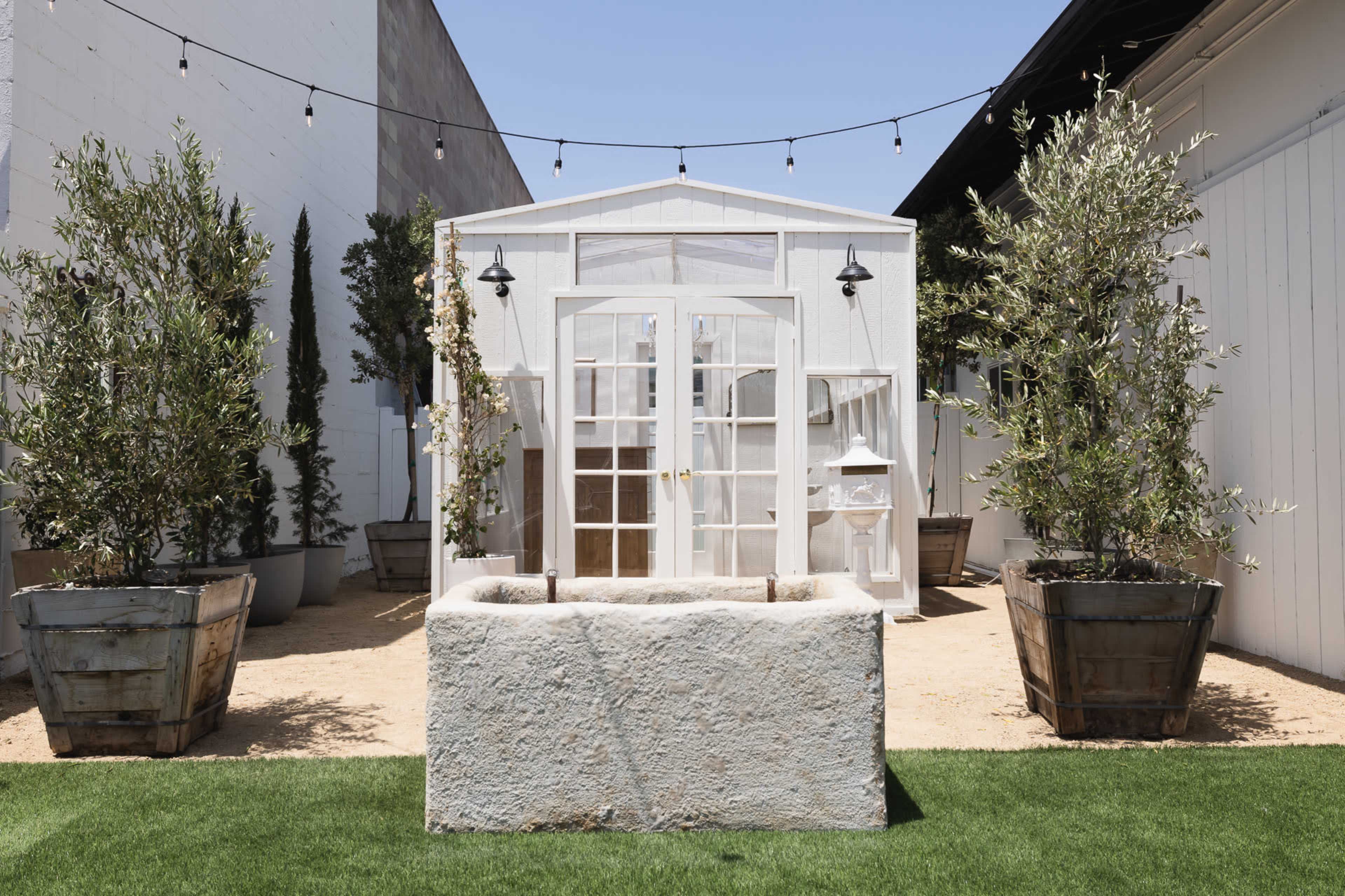 A white greenhouse with large glass doors is surrounded by potted olive trees and a stone planter in a sandy courtyard.