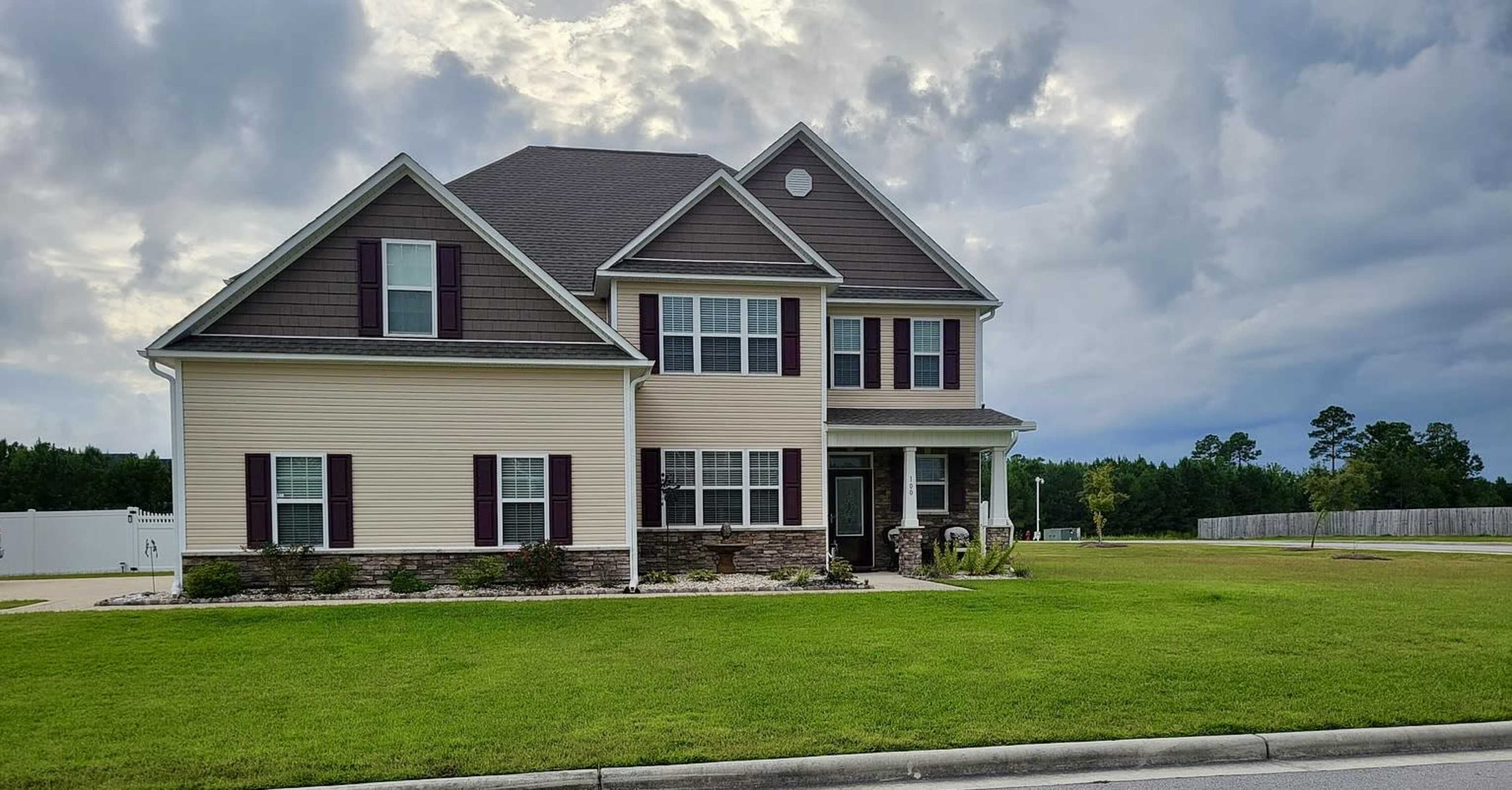 A two-story house with a combination of beige siding and dark brown accents sits on a green lawn under a cloudy sky.