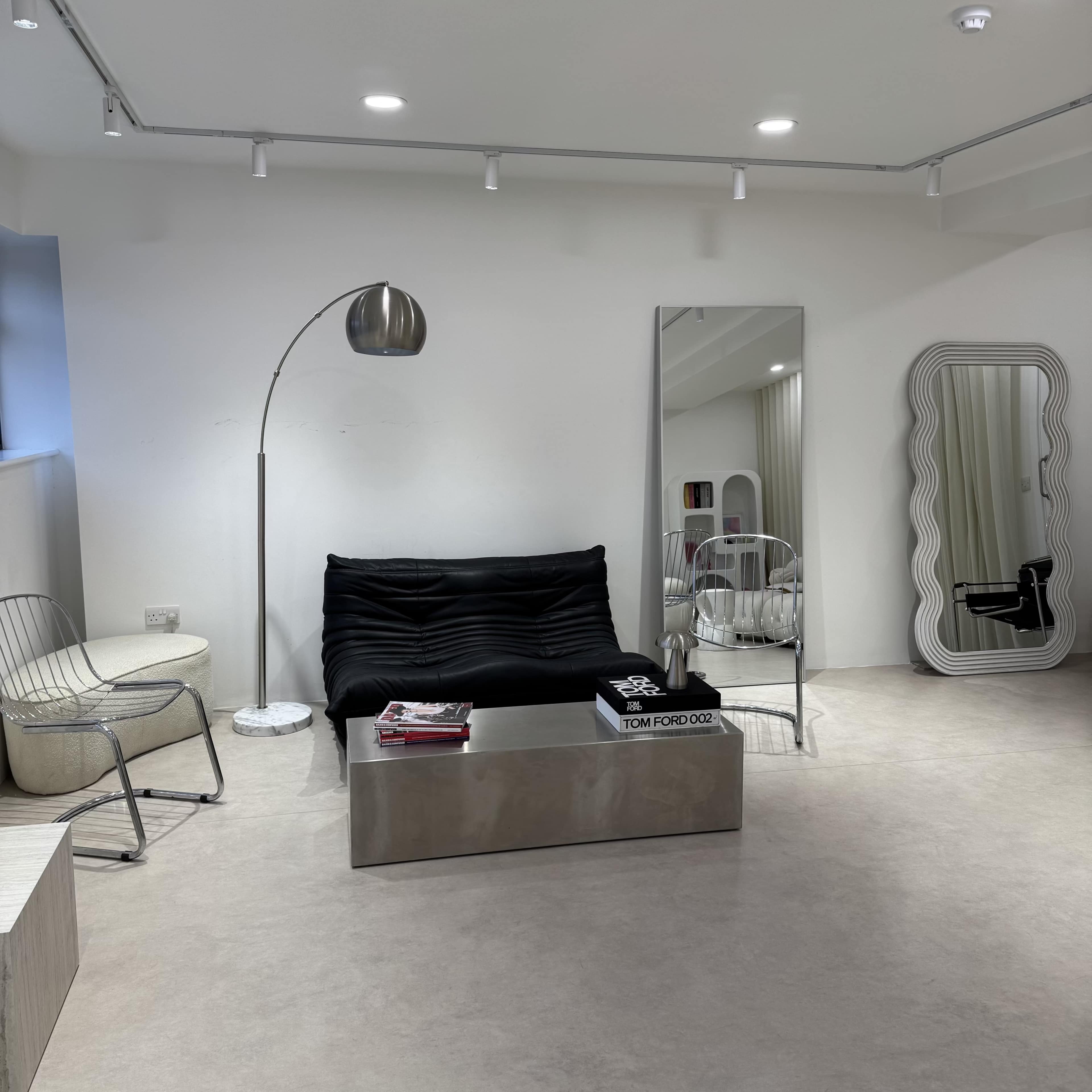 The image shows a modern interior with a black sofa, a silver coffee table, a floor lamp, and a large mirror, accompanied by minimalist furniture and decor elements.