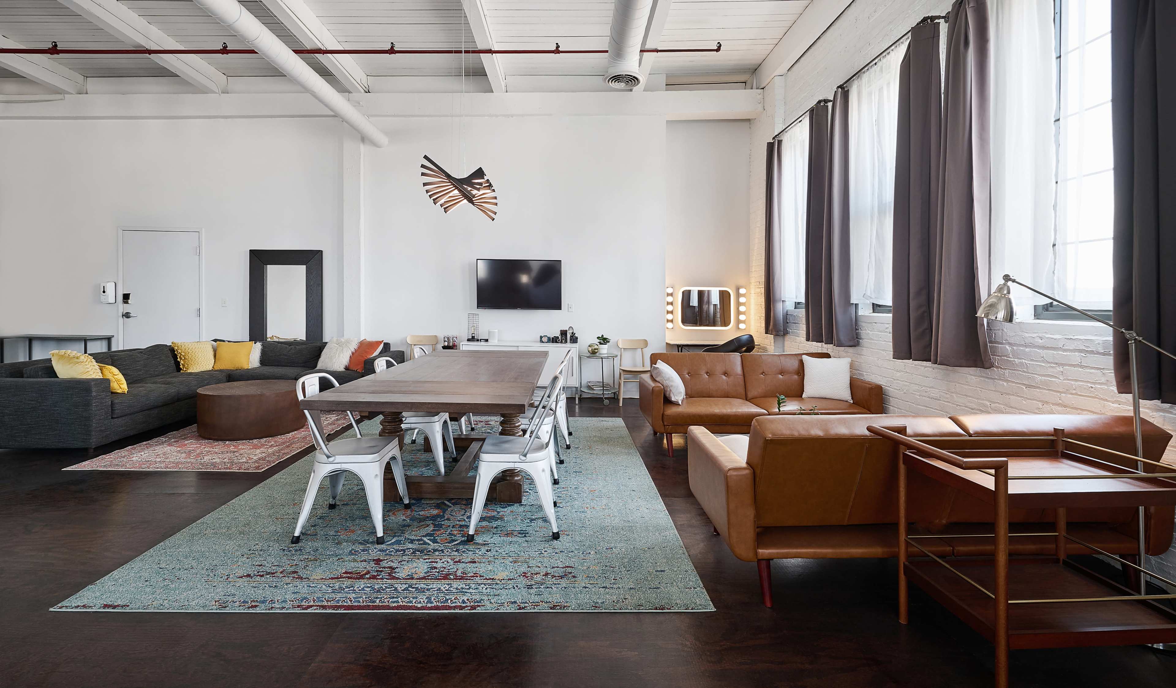 The image shows a modern loft-style living space featuring a large dining table, a sectional sofa, a coffee table, and large windows with curtains.