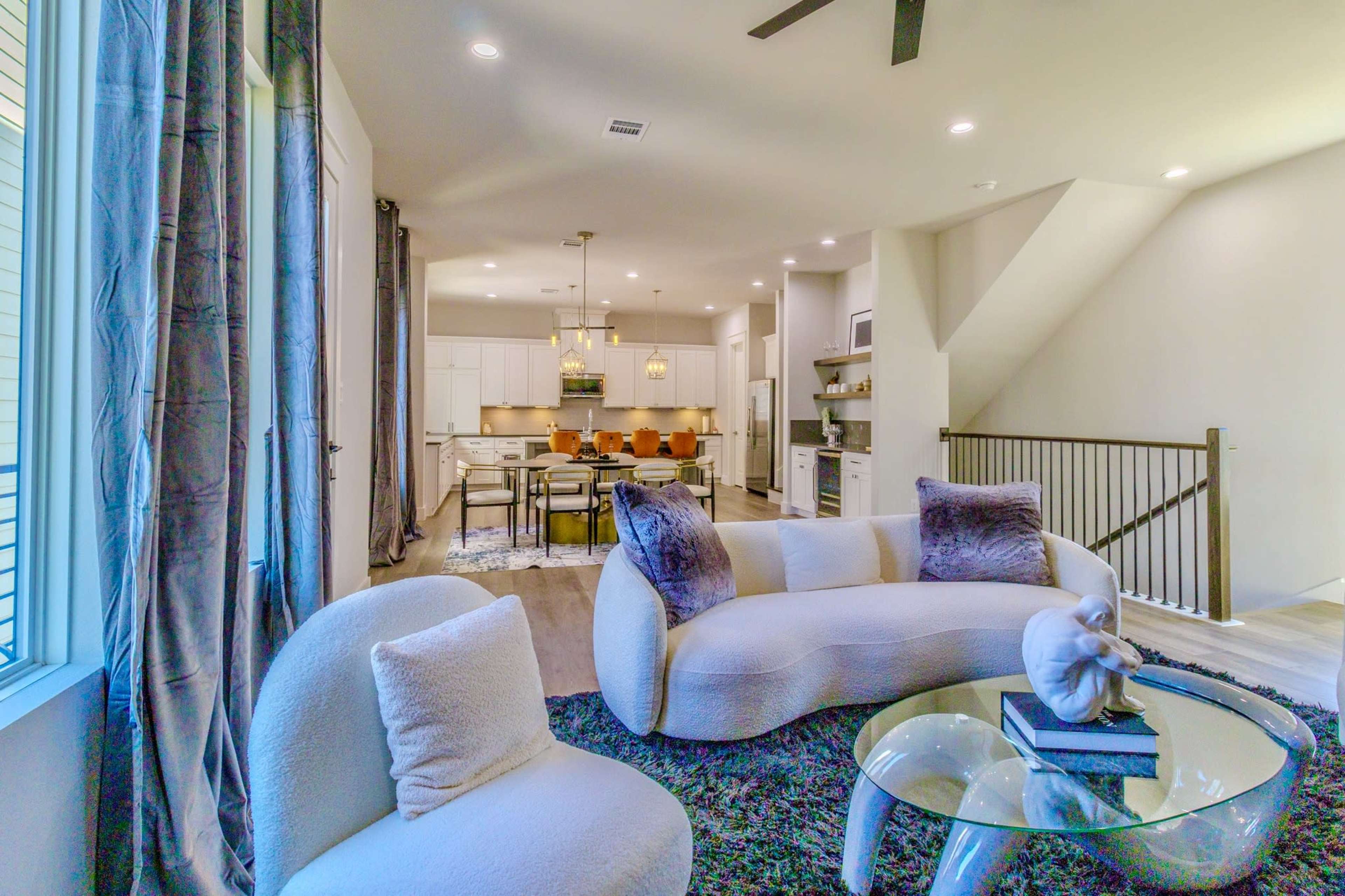 The image shows a modern living room that opens into a kitchen area, featuring a circular sofa, a glass coffee table, and a dining table with chairs in the background.