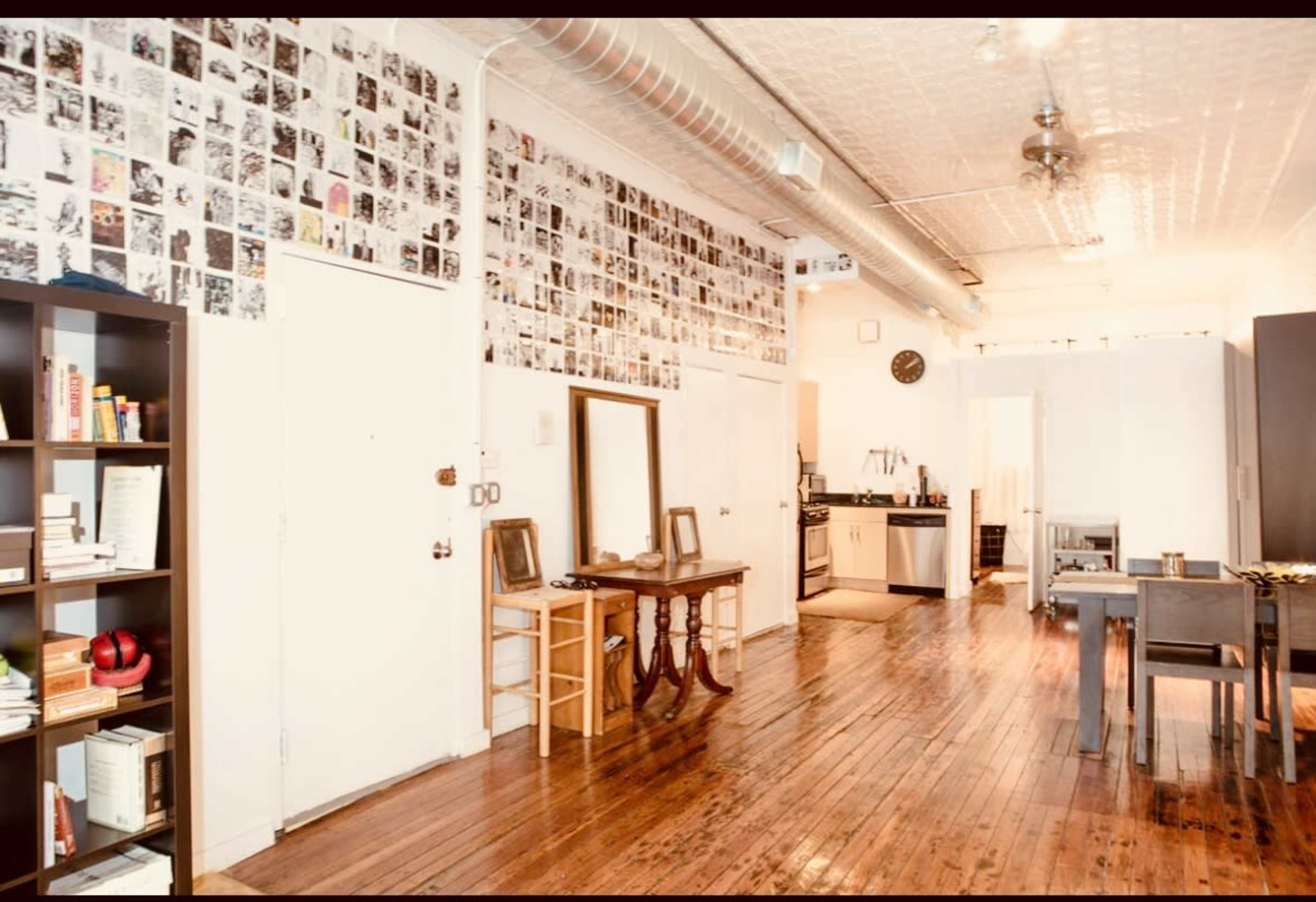 The image shows a spacious apartment interior with a wall covered in black-and-white photographs, wooden flooring, and various furniture pieces including a bookshelf, a dining table, and a mirror.