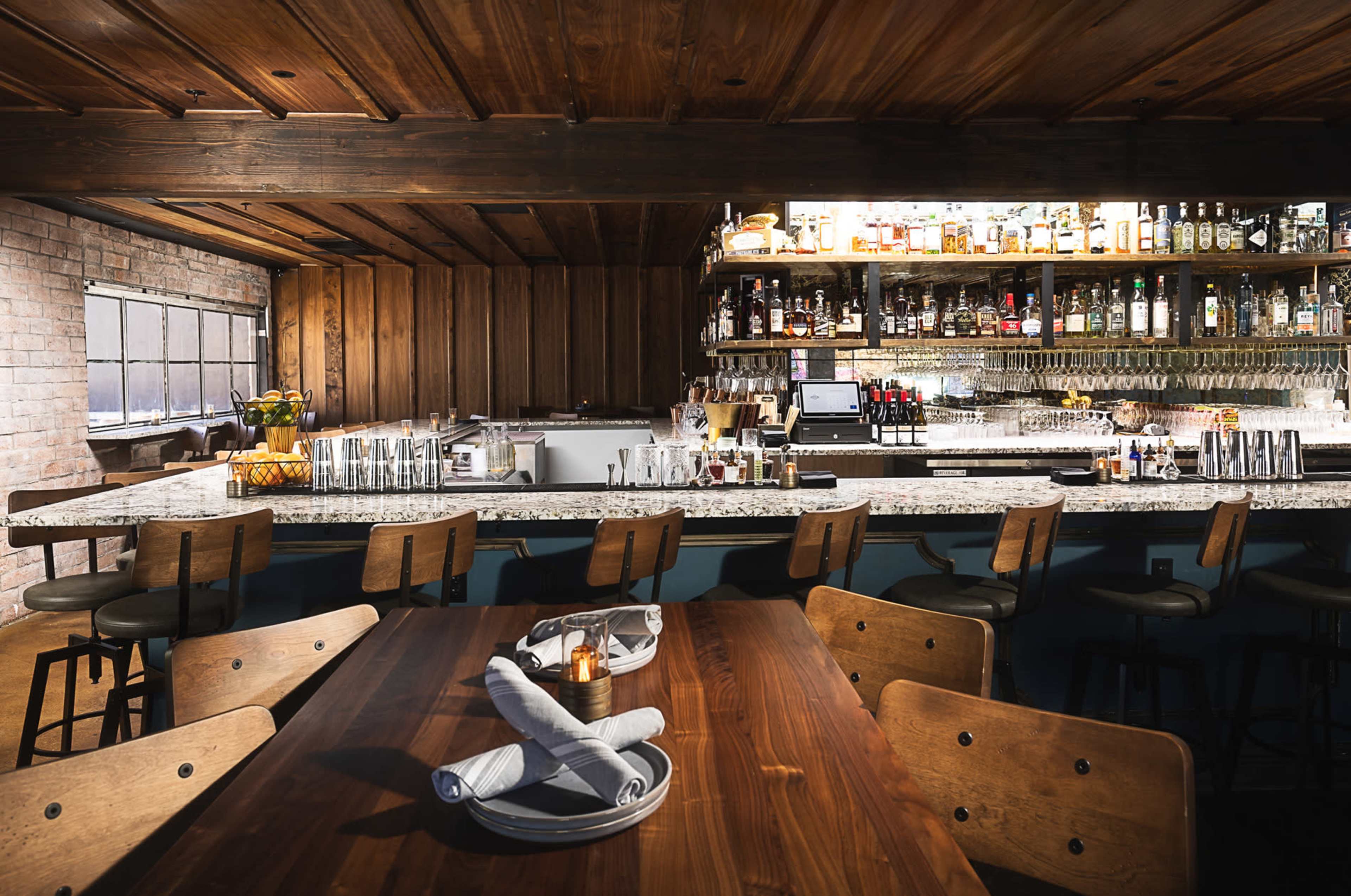 The image shows a rustic restaurant interior with wooden beams, a long bar displaying various bottles, and a dining table set for customers.