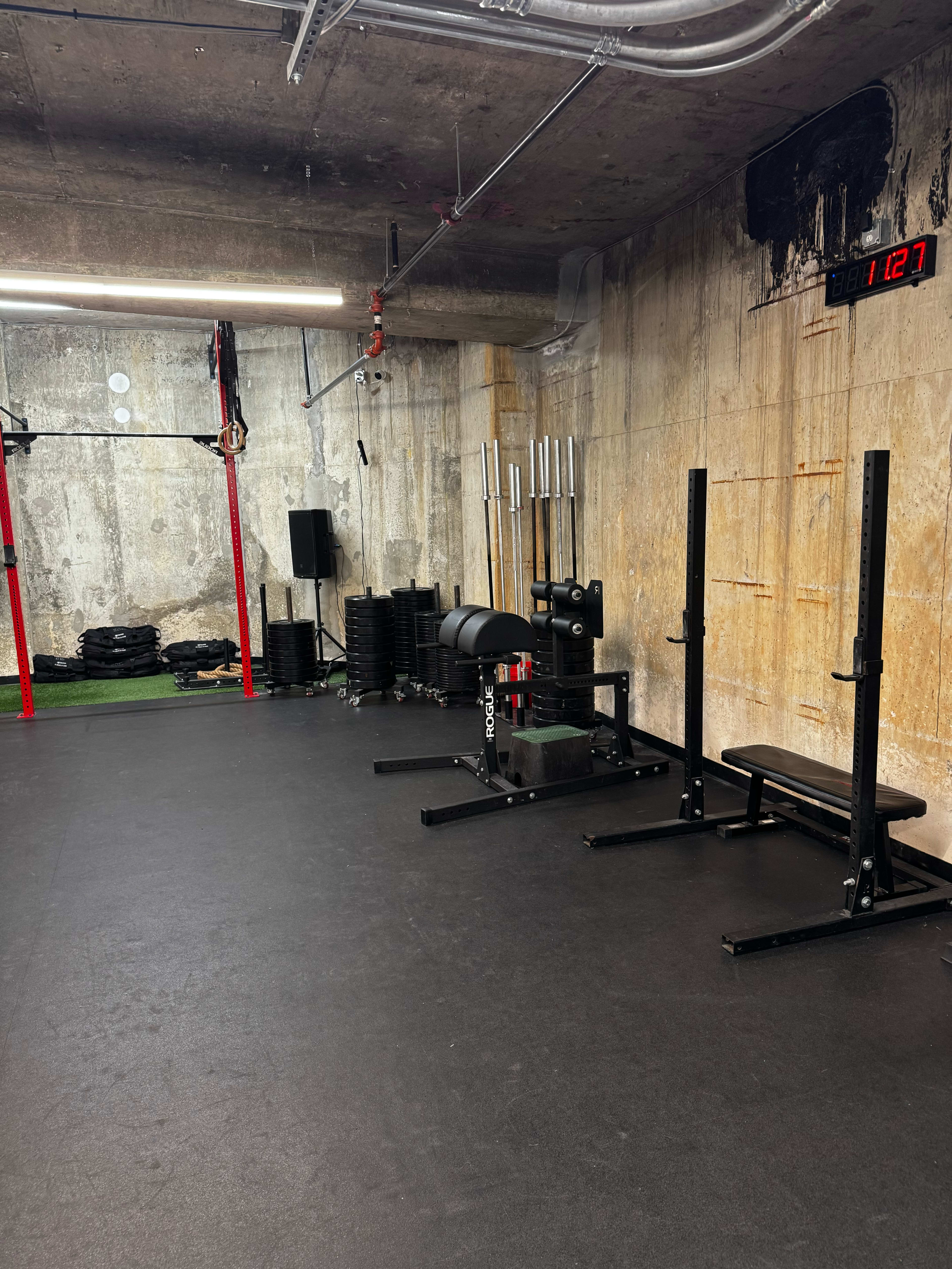The image shows a minimalist gym interior with weights, a bench, and exercise equipment against a concrete wall, featuring a digital clock on the right side.