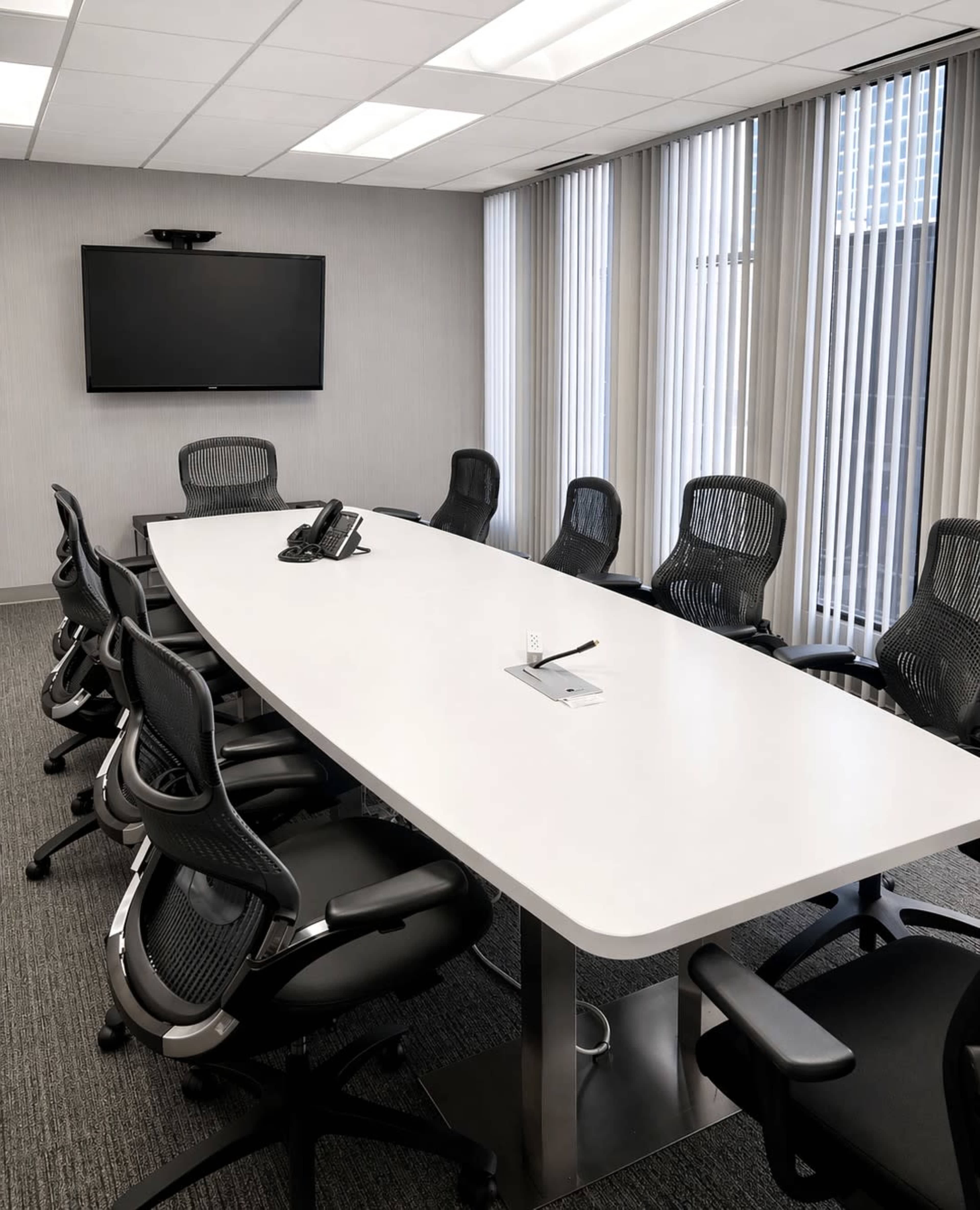 The image shows a modern conference room with a long, white table surrounded by black ergonomic chairs and a wall-mounted television.