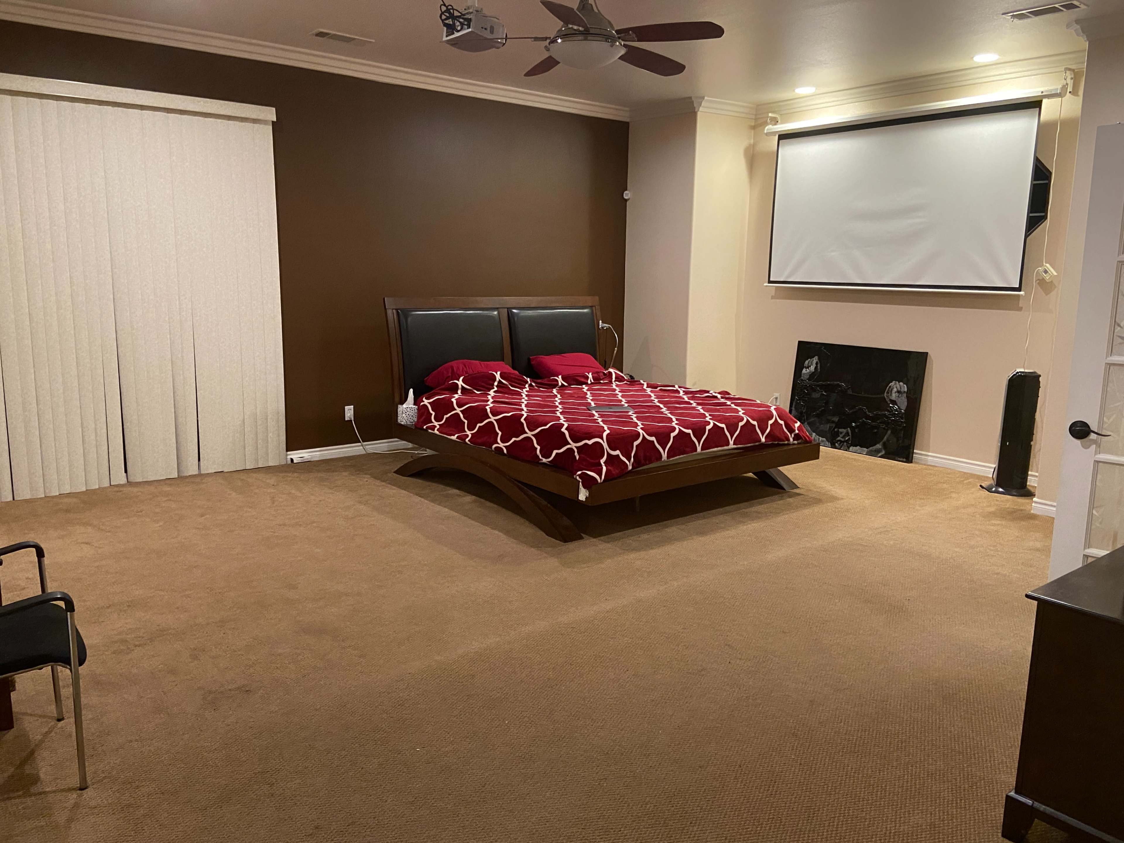 A spacious bedroom with a large bed featuring a red and white patterned comforter, a projector screen on the wall, and minimal furnishings.