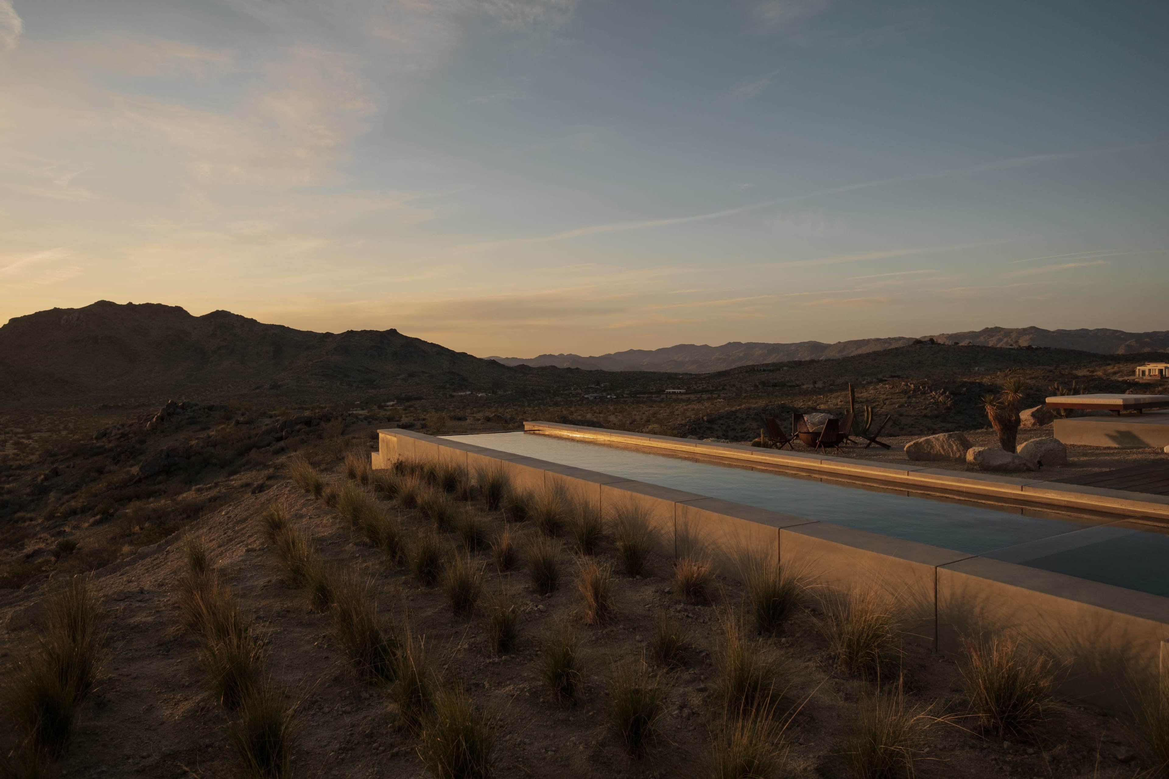 A modern infinity pool is positioned on a hillside with desert vegetation and mountains in the background during sunset.