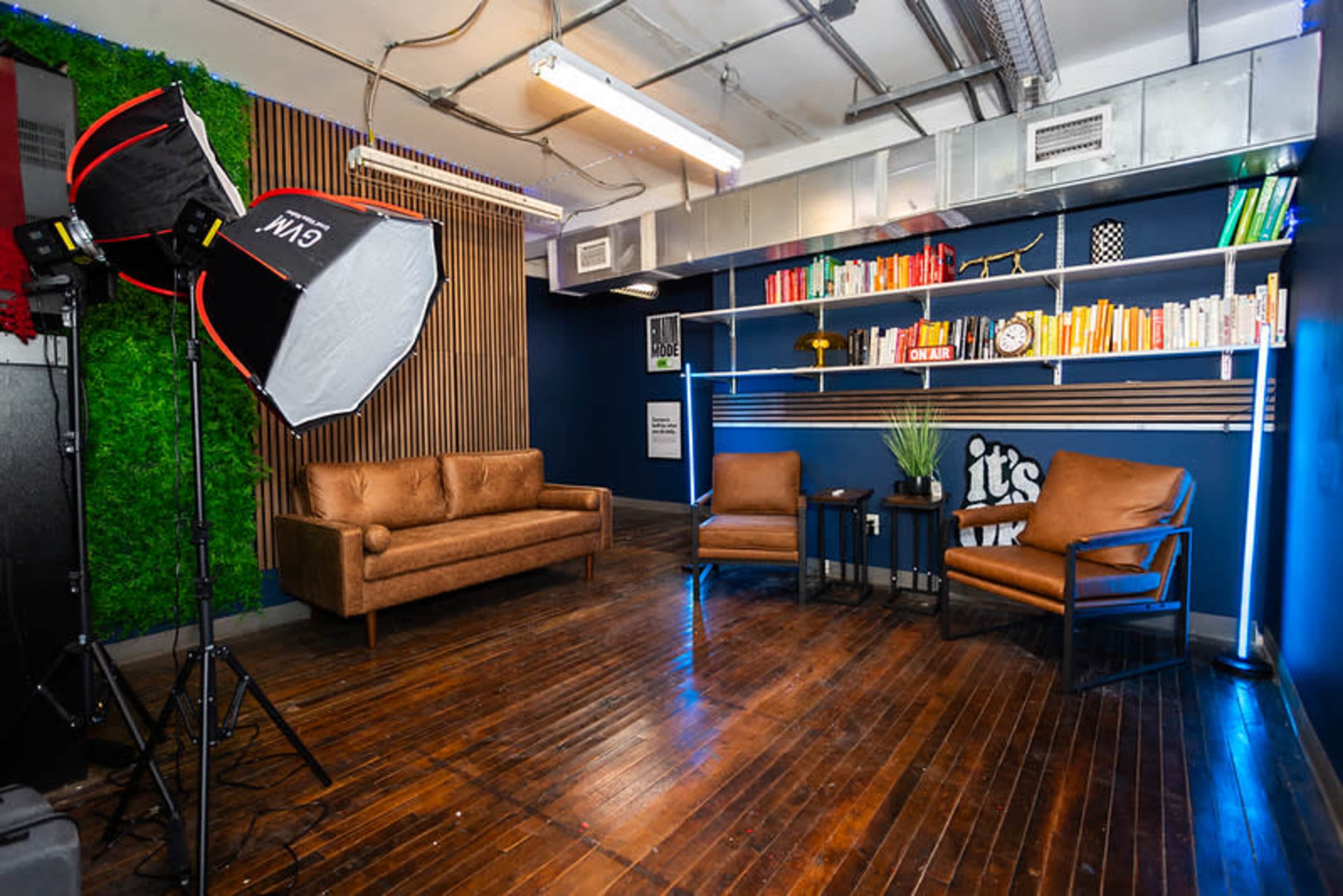 The image shows a modern studio space with a brown leather sofa, two chairs, a photography light setup, and shelves filled with books against a green wall.