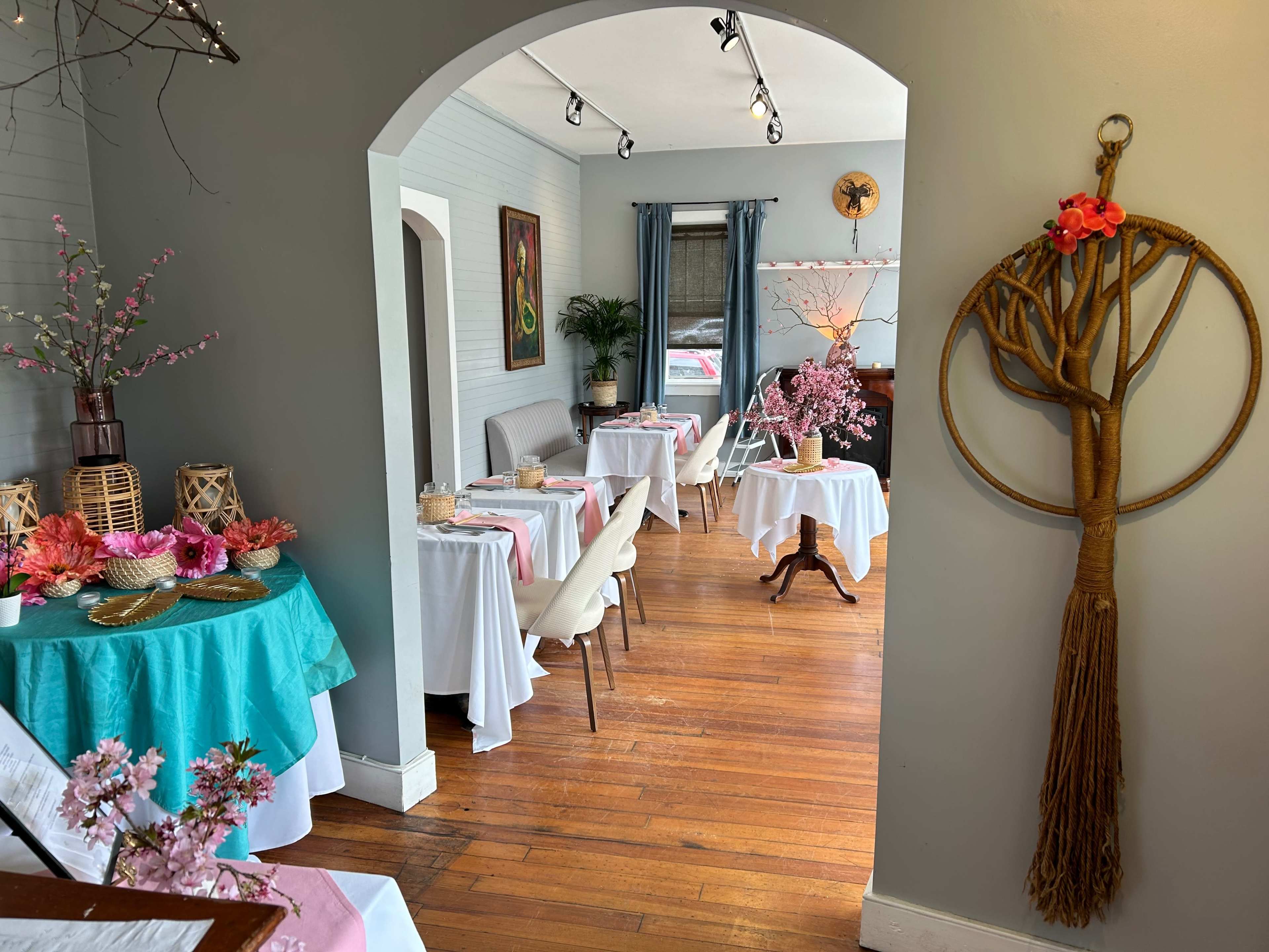 A dining area with tables dressed in white and turquoise tablecloths, adorned with pink floral arrangements, and a decorative wall hanging beside an entrance.