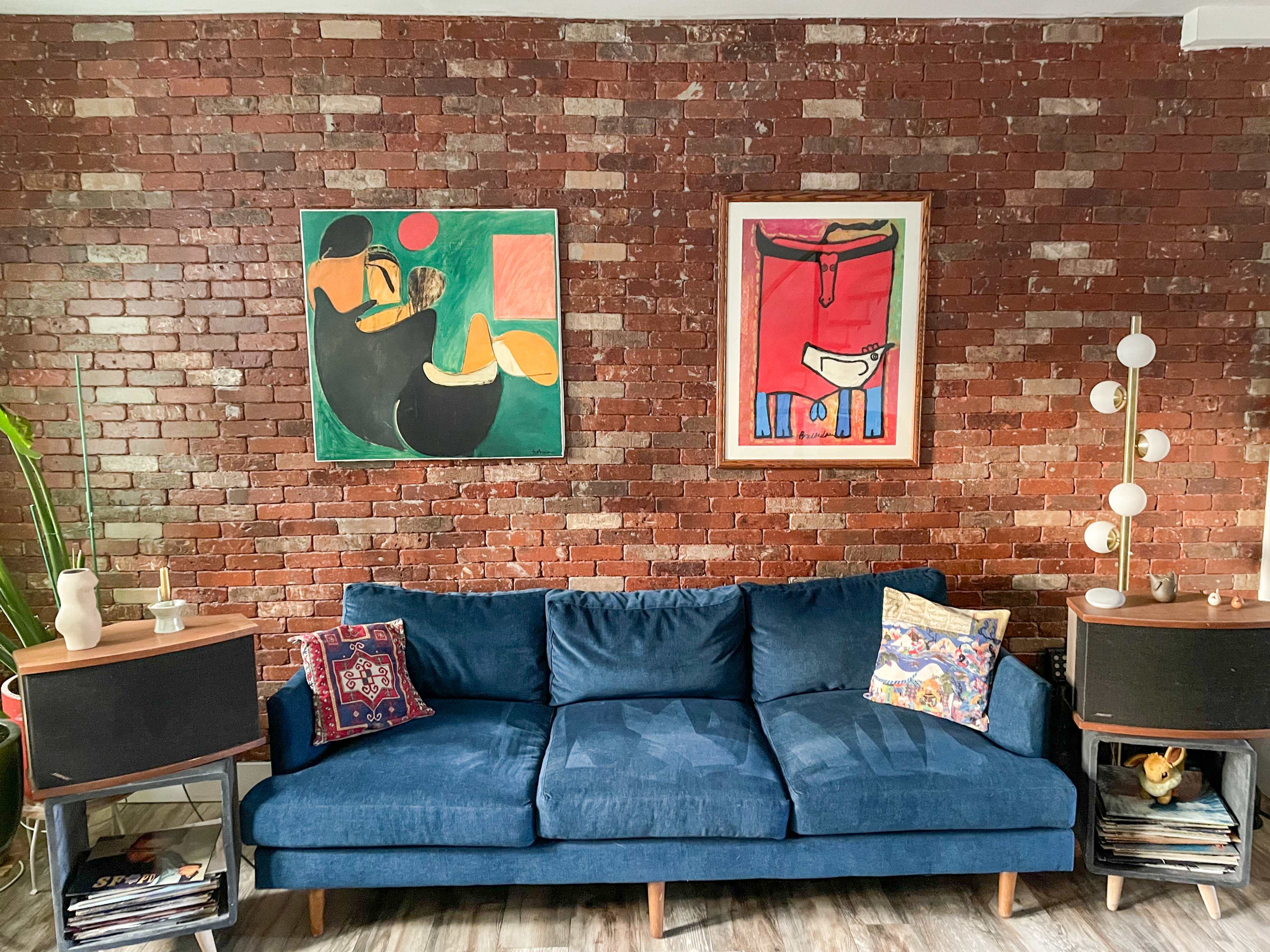 The image shows a blue couch against a brick wall, with two framed art pieces above it and side tables displaying decorative objects.