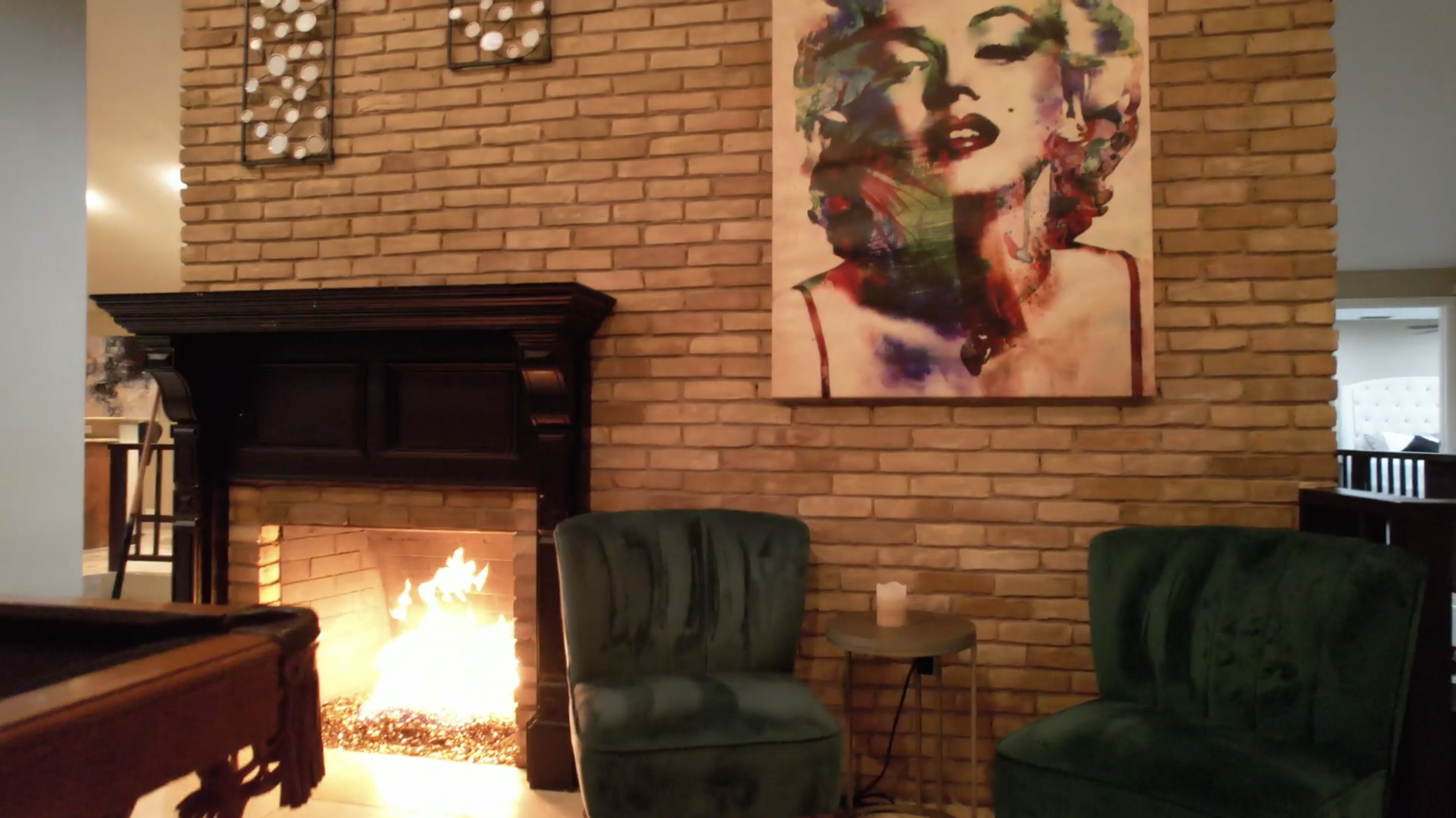 A cozy room features a brick wall with an abstract painting of a woman above a fireplace, flanked by two green armchairs and a small table.