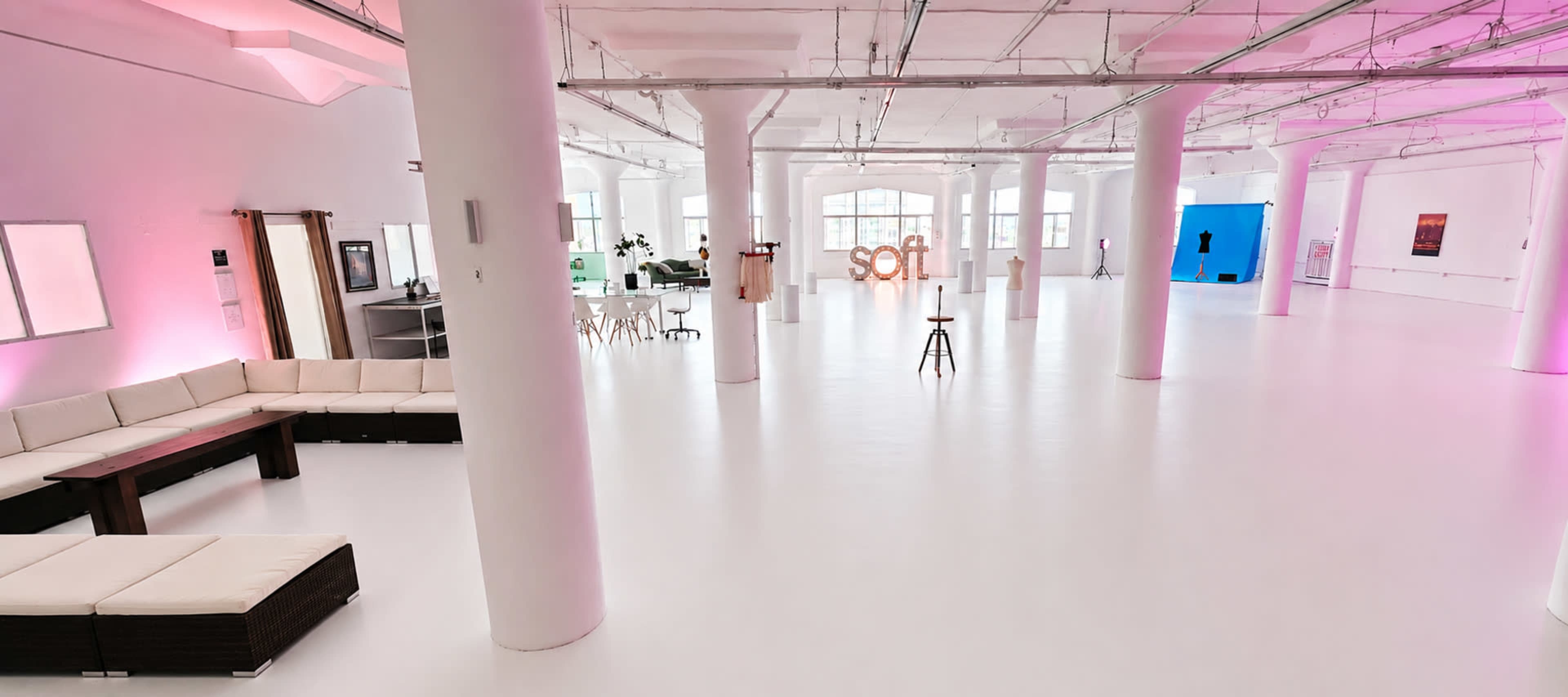 The image shows a spacious, modern interior with white flooring, multiple columns, and soft pink lighting, featuring lounge seating and a decorative "soft" sign in the center.