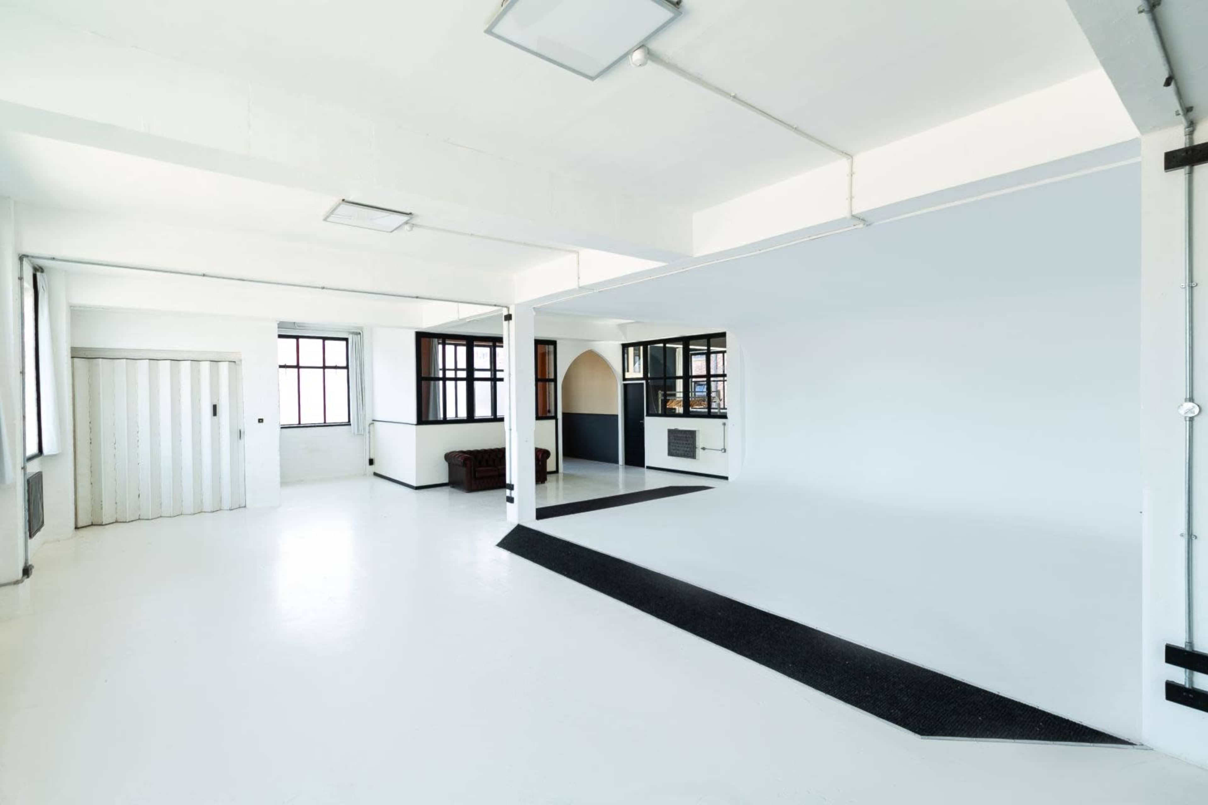 A spacious, empty interior with white walls, a polished floor, and large windows, featuring a small partitioned area in the center.