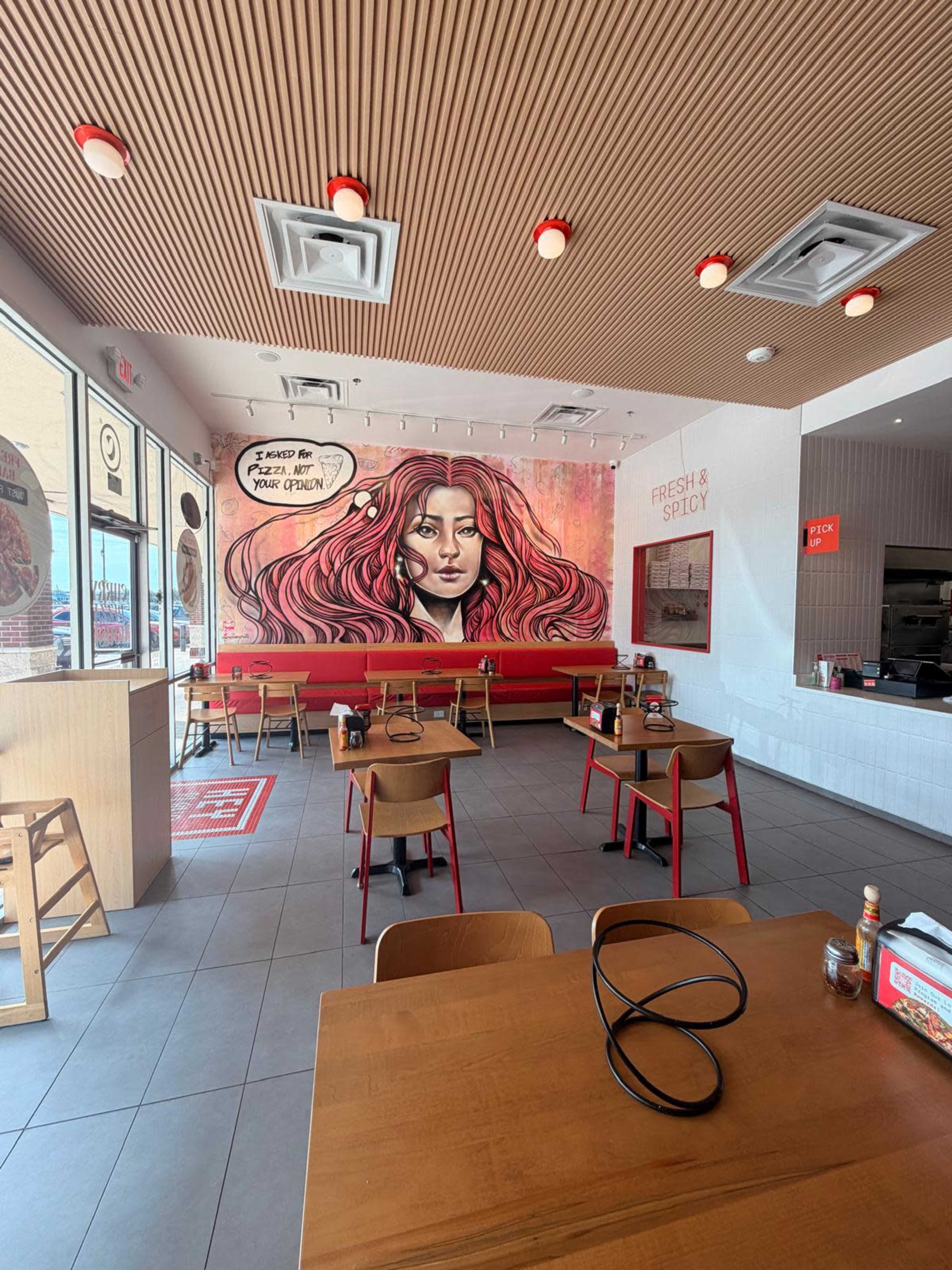 The interior of a fast-casual restaurant features wooden tables and chairs, with a large mural of a woman on the wall and bright red accents.