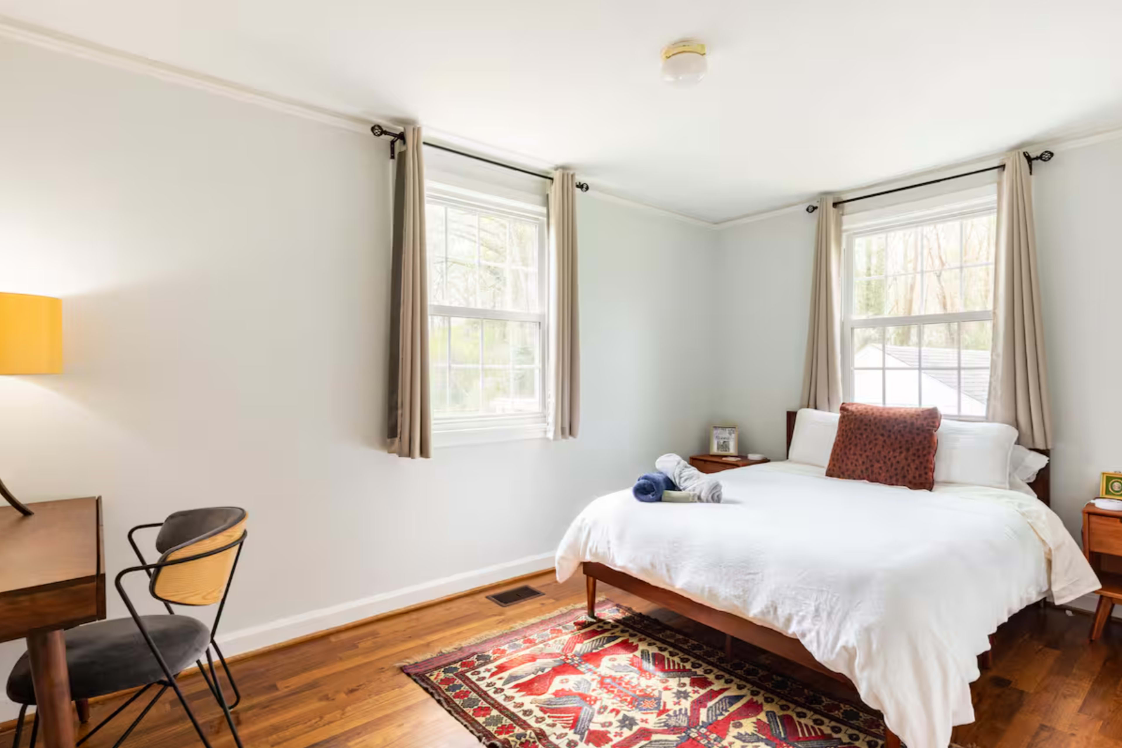 The image shows a minimalist bedroom featuring a double bed with white linens, a desk with a chair, and a colorful area rug on wooden flooring.