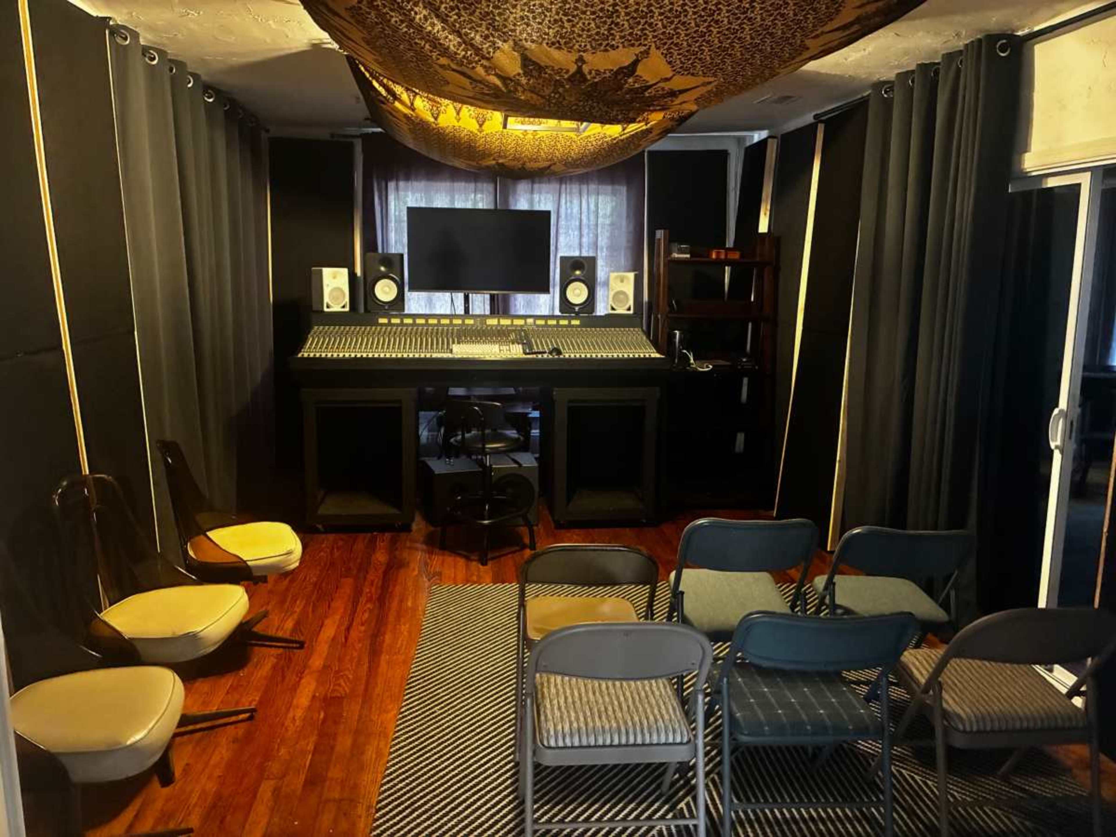 The image shows a music studio with a mixing console, speakers, a computer monitor, and seating arranged in front.