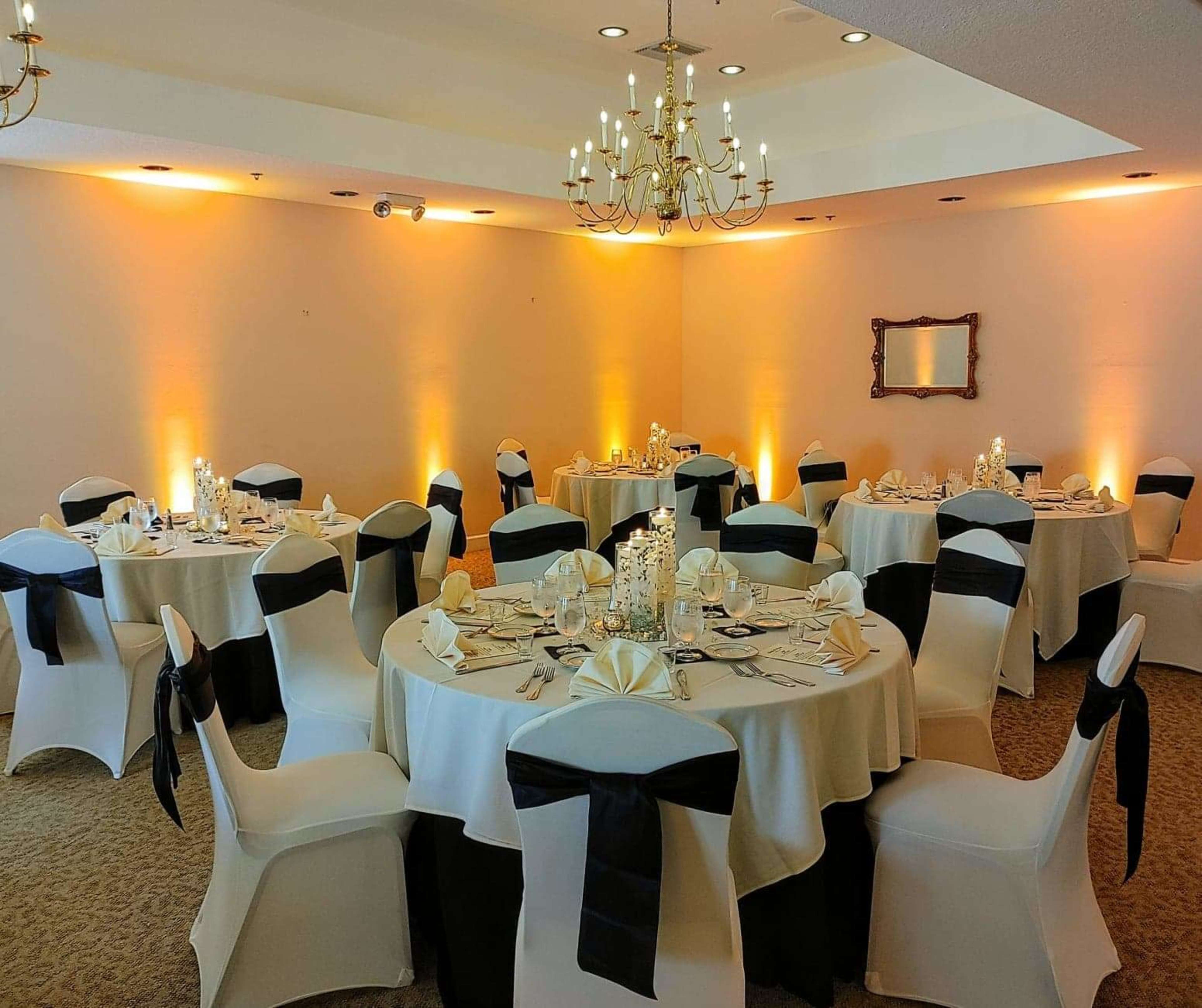 A decorated dining area with round tables covered in white tablecloths and black sashes, surrounded by chairs, arranged for an event.