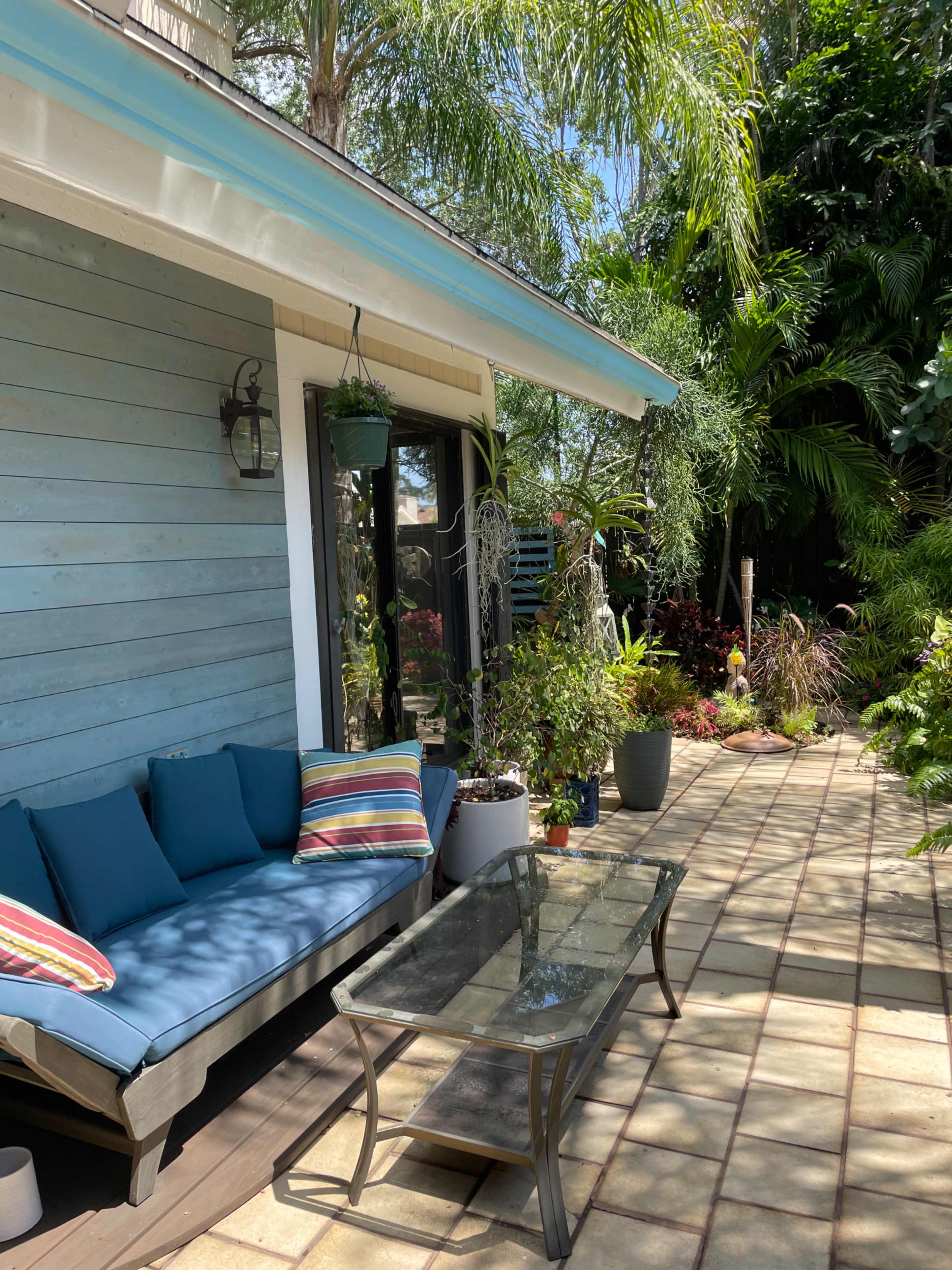 A patio with a couch, striped cushions, and a glass coffee table is surrounded by lush greenery and potted plants.