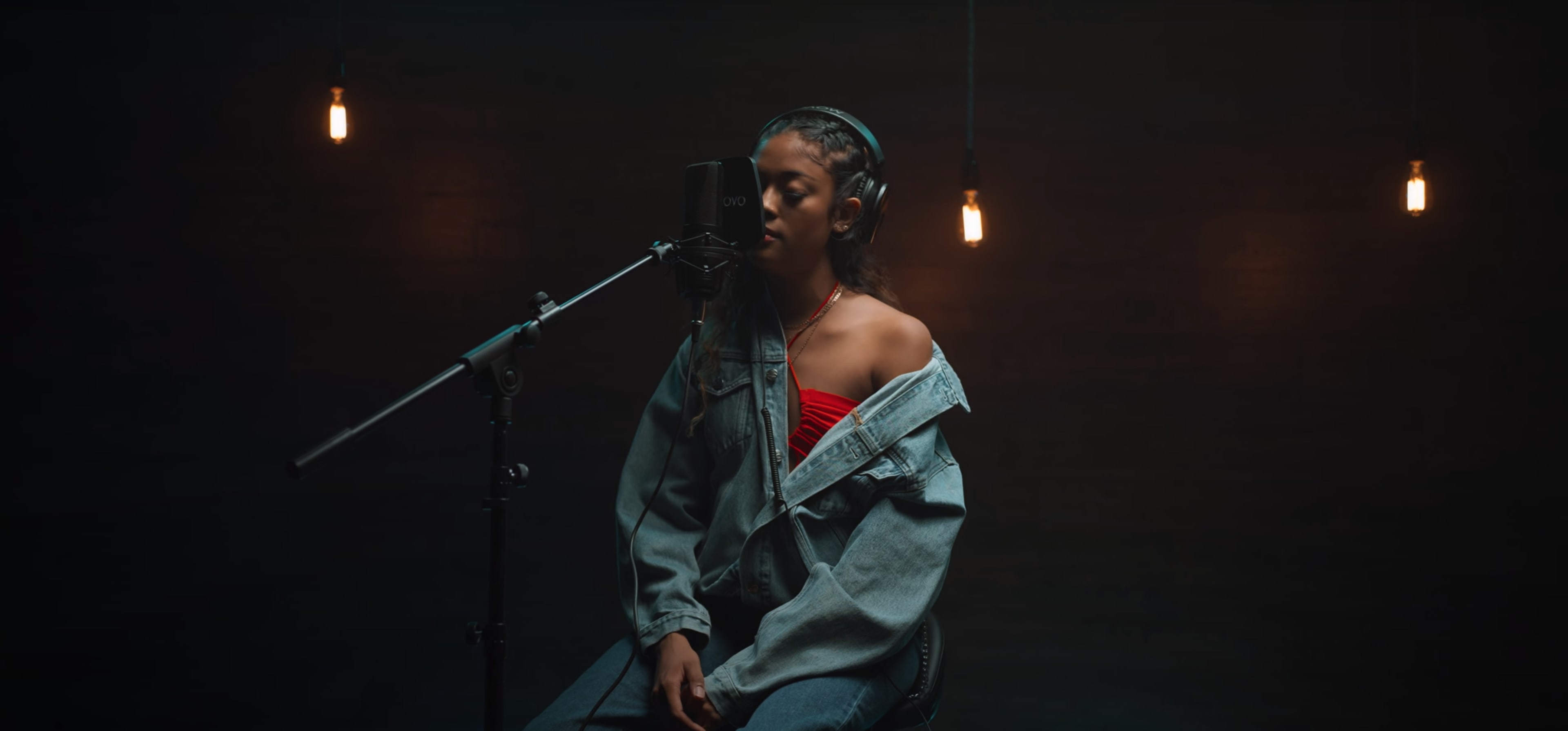 A woman sits in front of a microphone wearing a red top and a denim jacket, with headphones on, in a dimly lit recording studio.