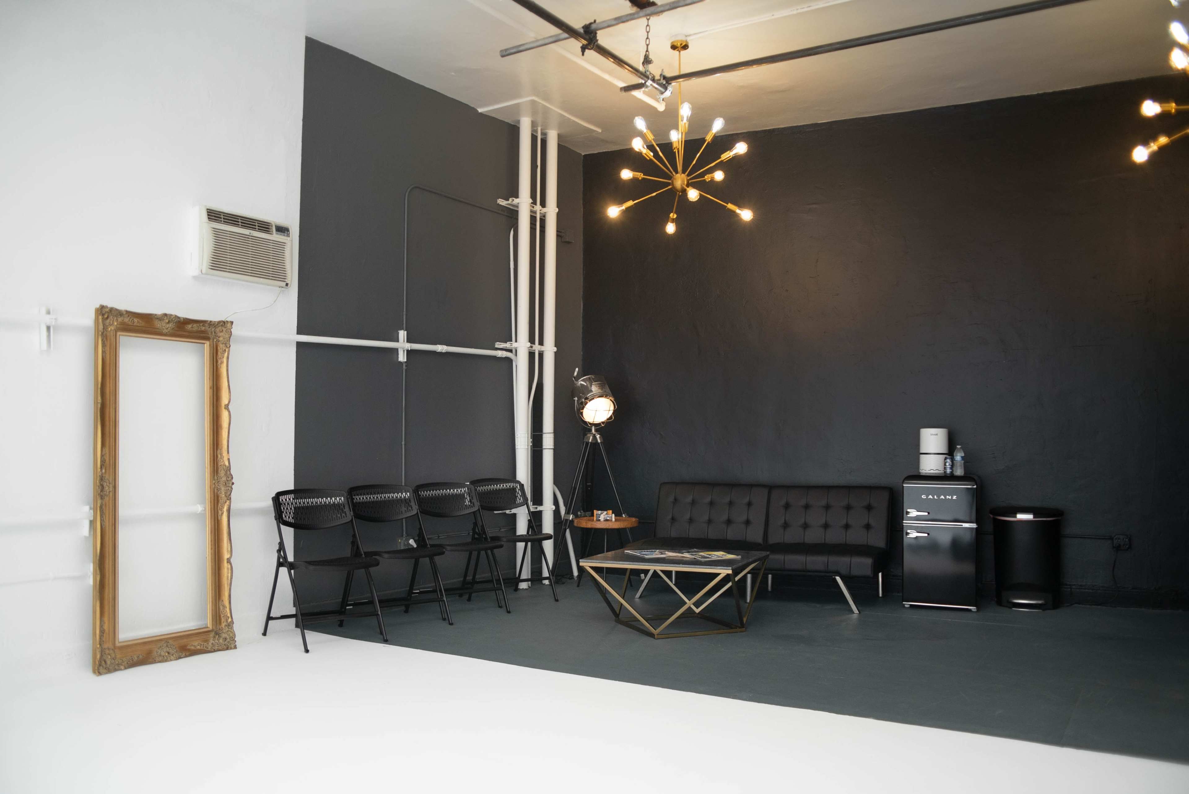 A minimalist interior space featuring a black wall, a tufted black sofa, a gold starburst chandelier, a small refrigerator, and a round glass coffee table surrounded by black chairs.
