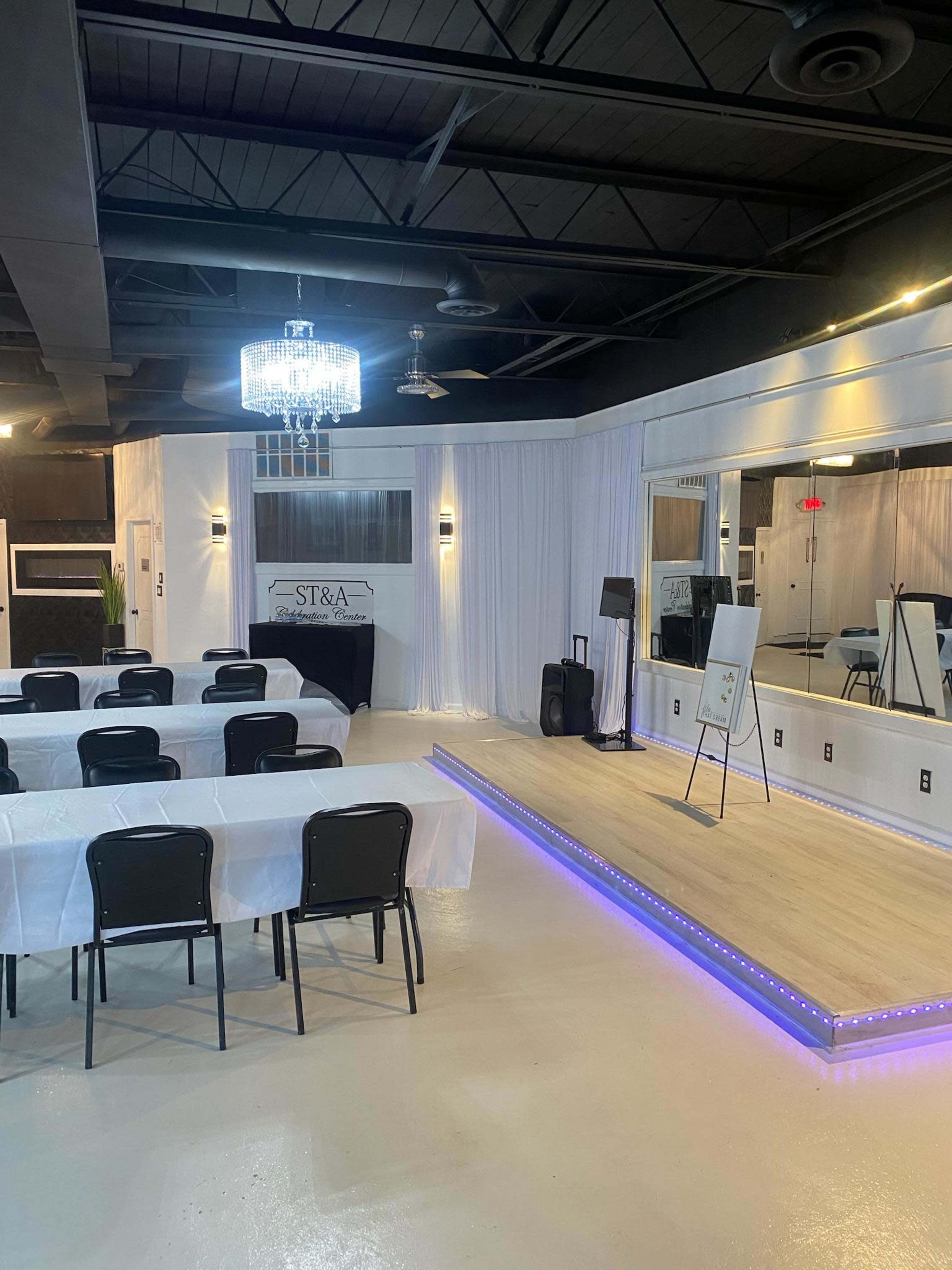 The image shows an event space featuring white tablecloths on several tables, a stage with a lit edge, a chandelier, and a mirrored wall.