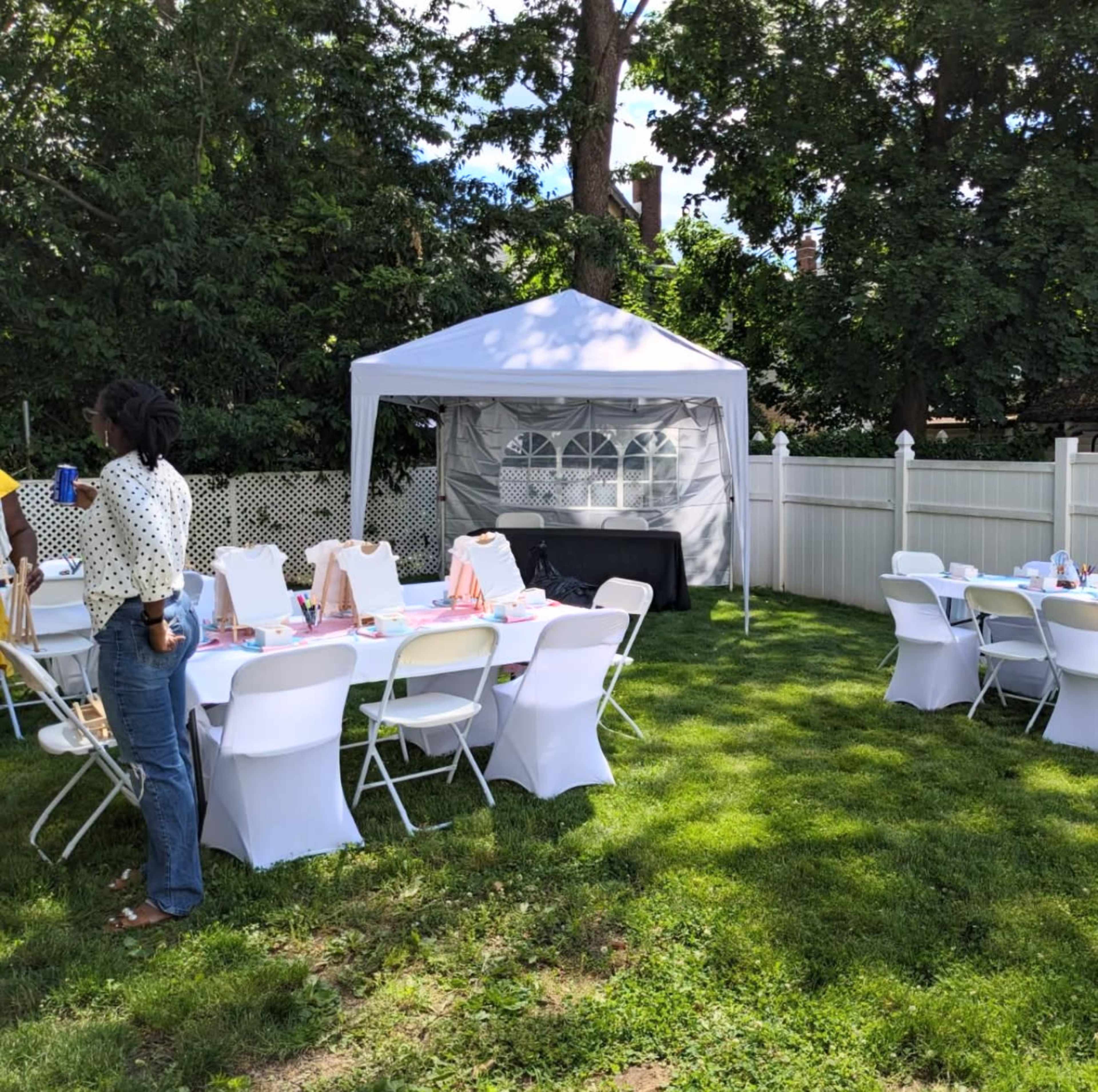 A backyard is set up for an outdoor gathering, featuring a white tent and several tables with chairs arranged on green grass.