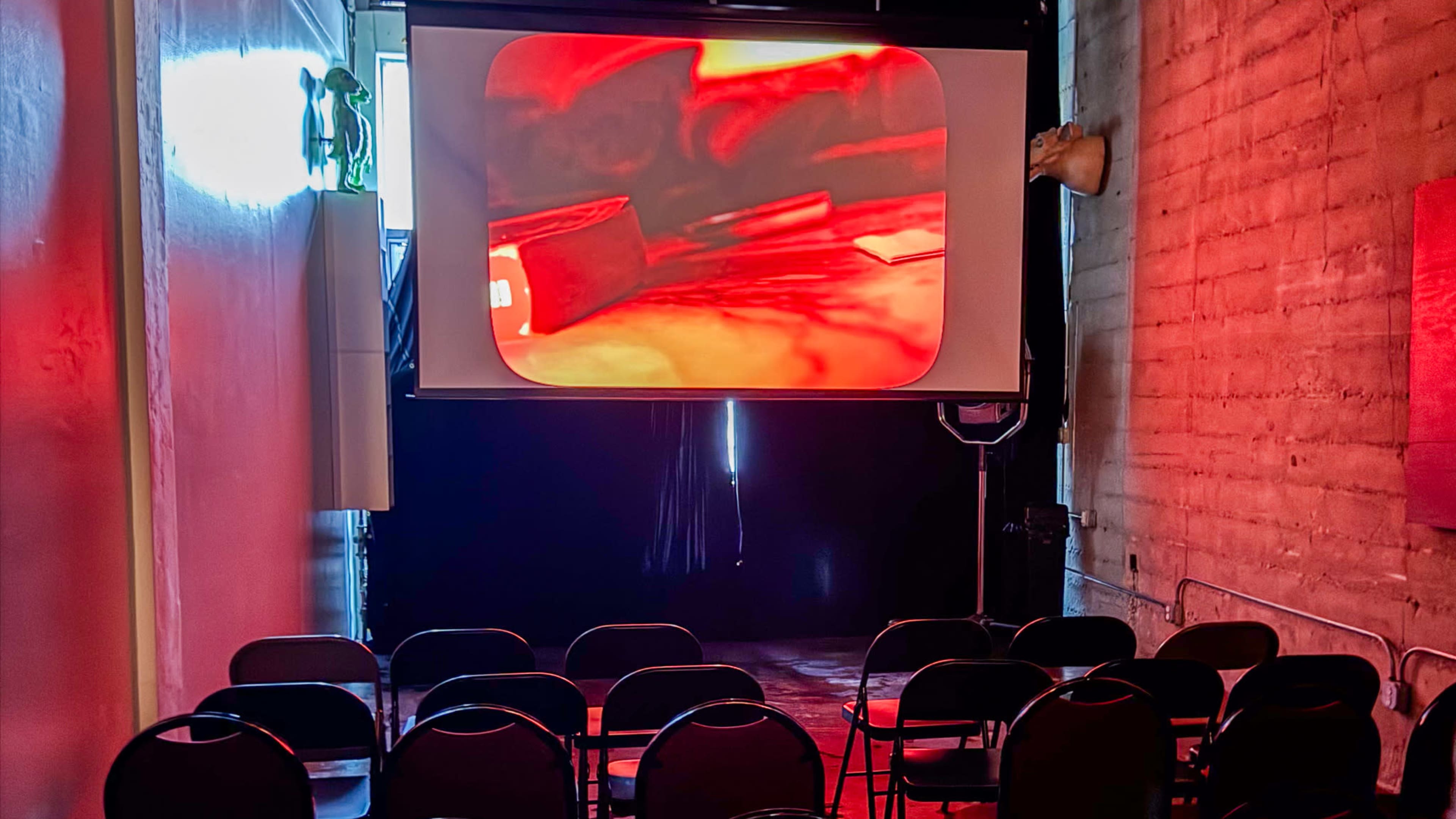 A projection screen displays a red-tinted image in a dimly lit room with empty black chairs arranged in front.