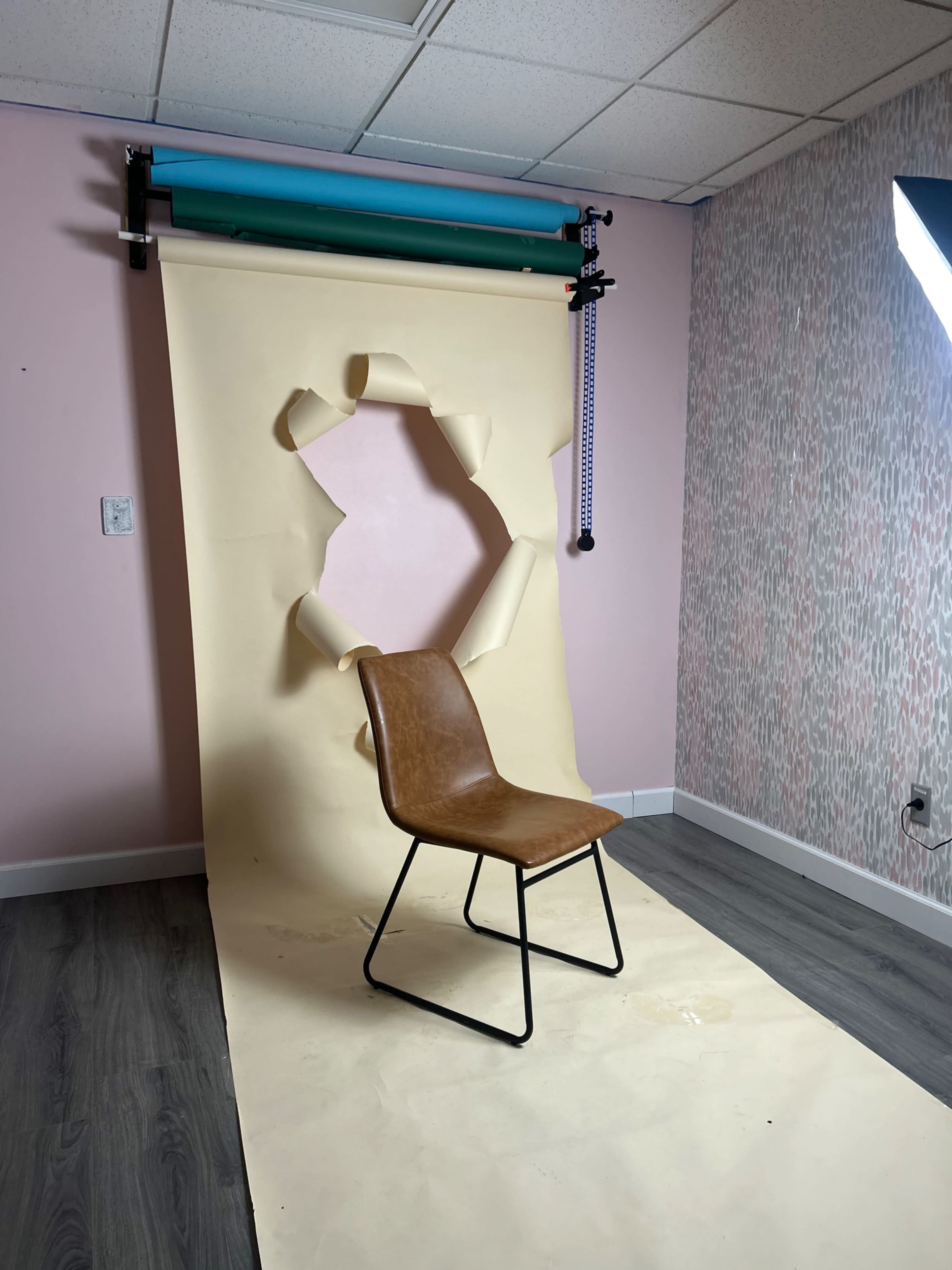 A brown chair is positioned in front of a torn backdrop against a pink wall with patterned wallpaper.