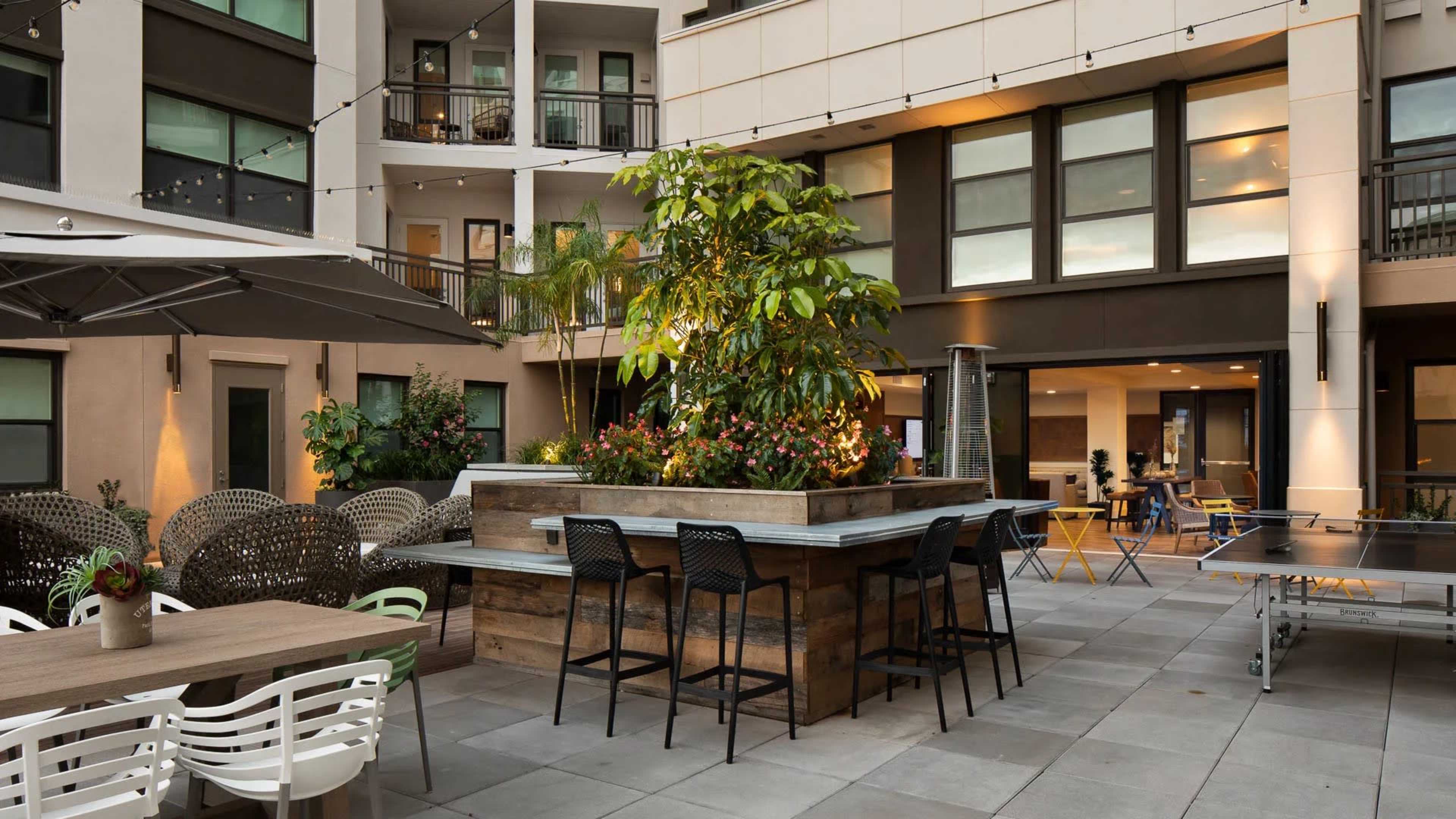 A modern outdoor courtyard with a communal seating area, a bar-style table, and lush greenery surrounded by buildings.
