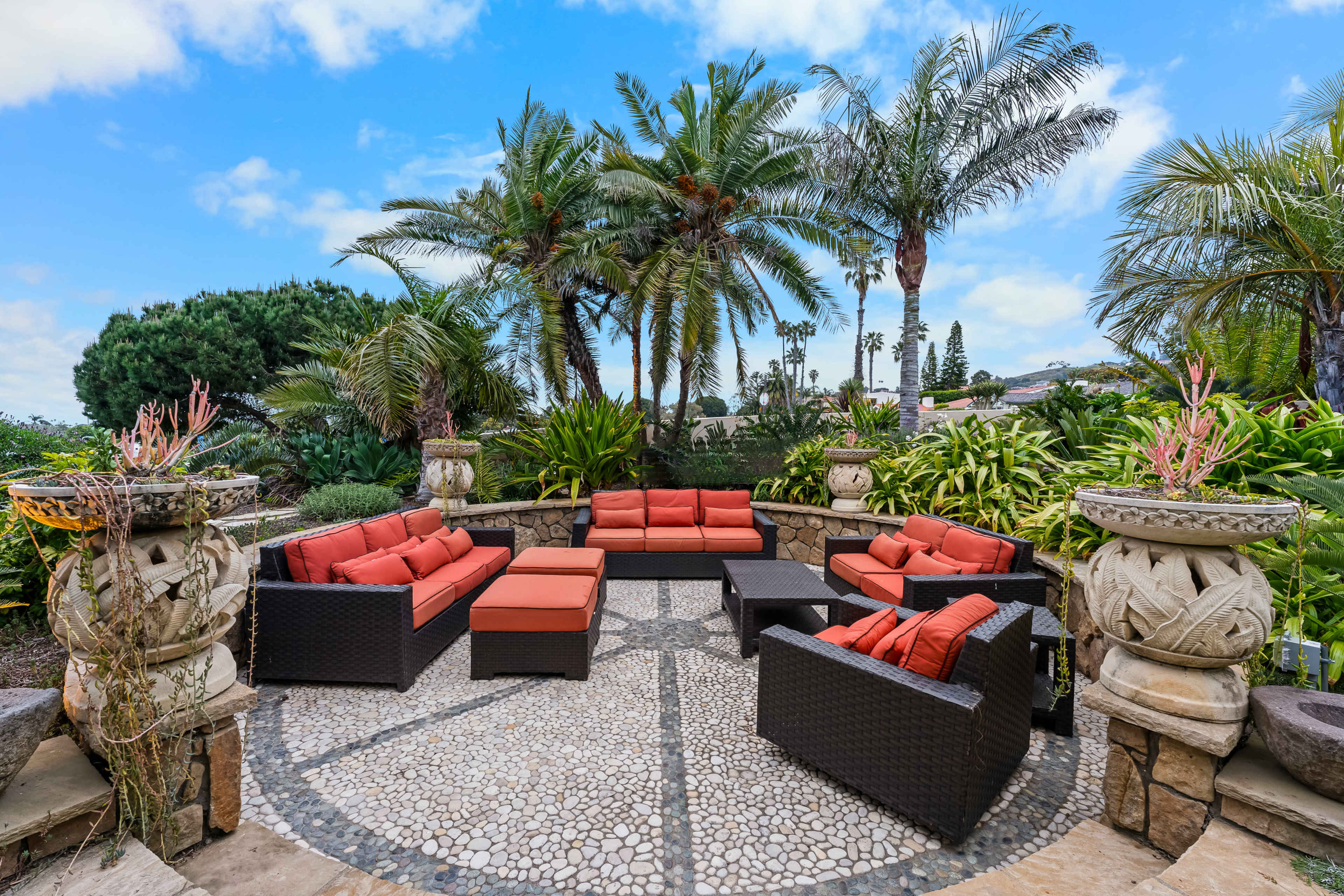 A landscaped patio features a circular stone area surrounded by palm trees and shrubs, furnished with a set of red cushioned outdoor sofas and a central coffee table.