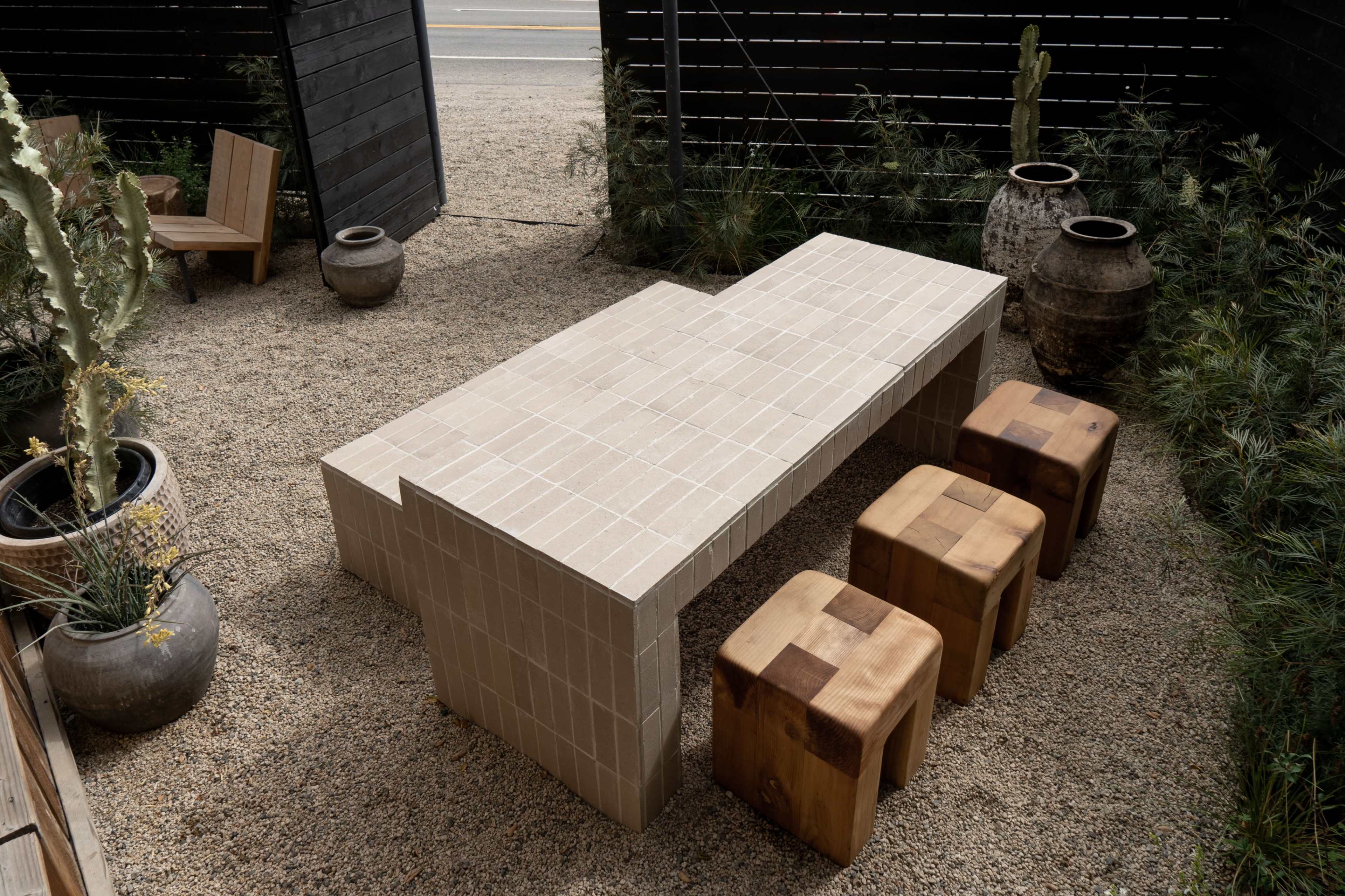 The image shows a long, rectangular concrete table with wooden stools in a gravel outdoor setting surrounded by potted plants and a wooden fence.