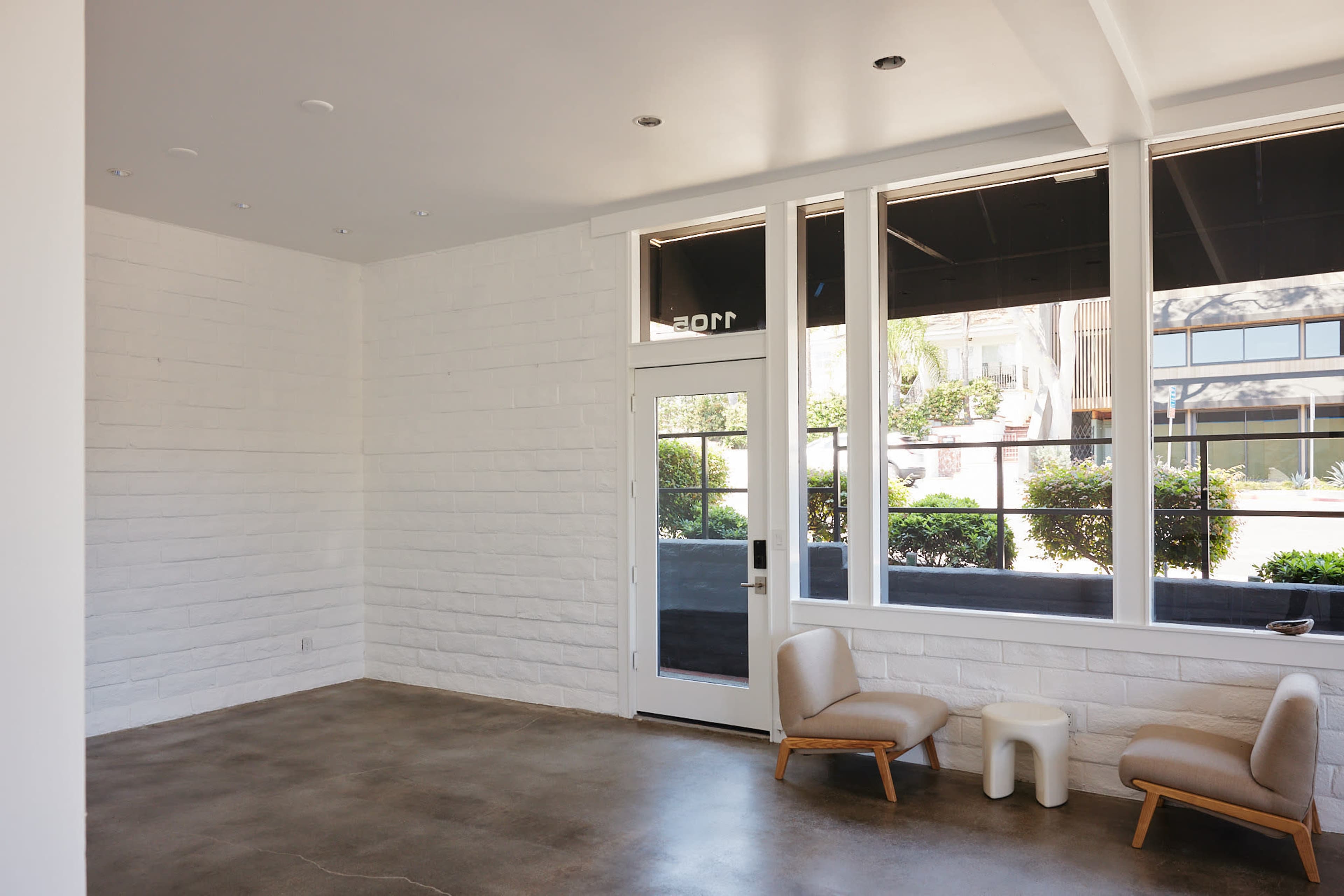 The image shows a minimalist interior of a commercial space featuring a white brick wall, large windows, and two light-colored chairs on a concrete floor.