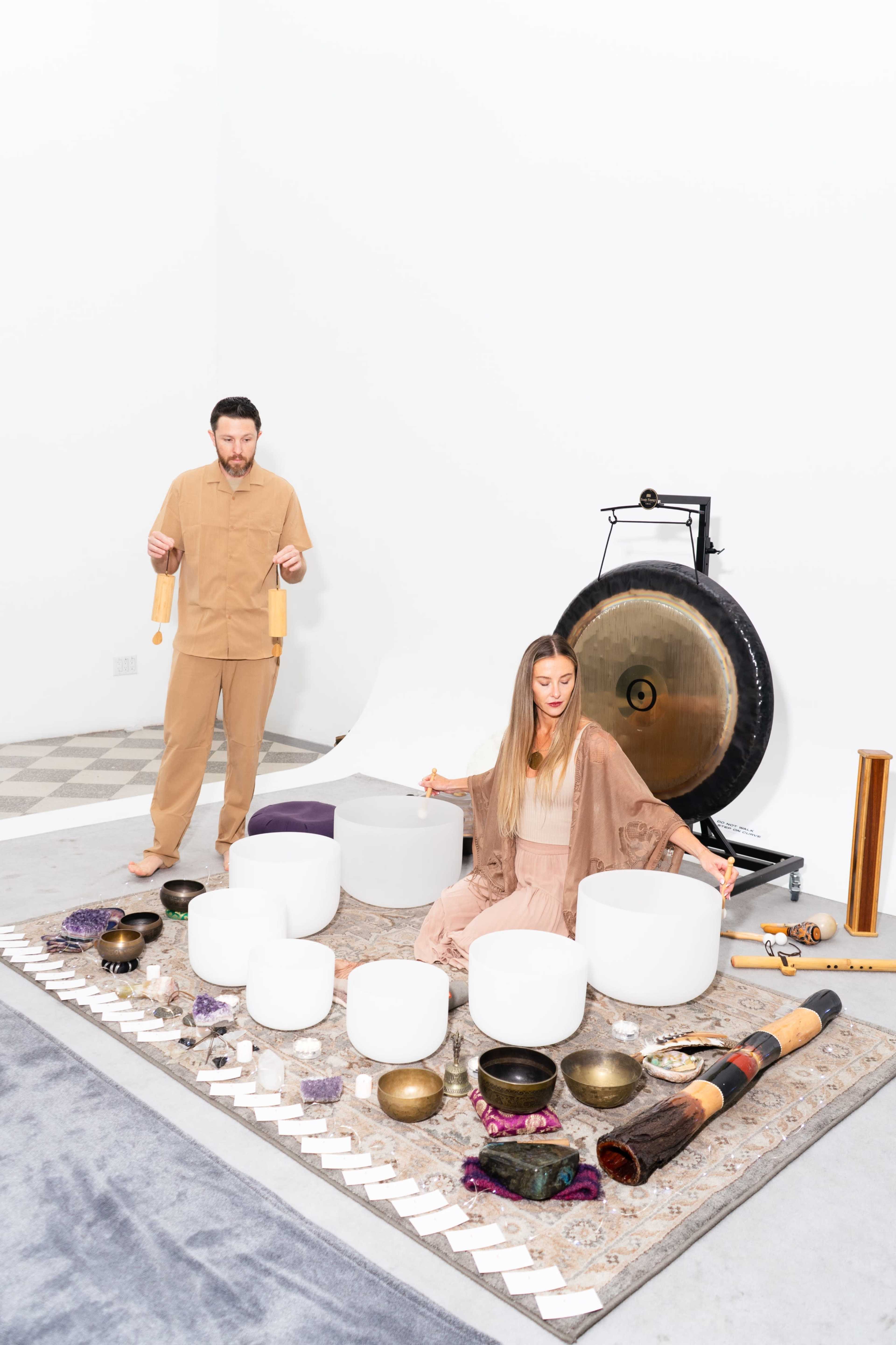 A man and a woman are seated on a patterned rug surrounded by various musical instruments, including crystal bowls and a gong, in a minimalist room with white walls.