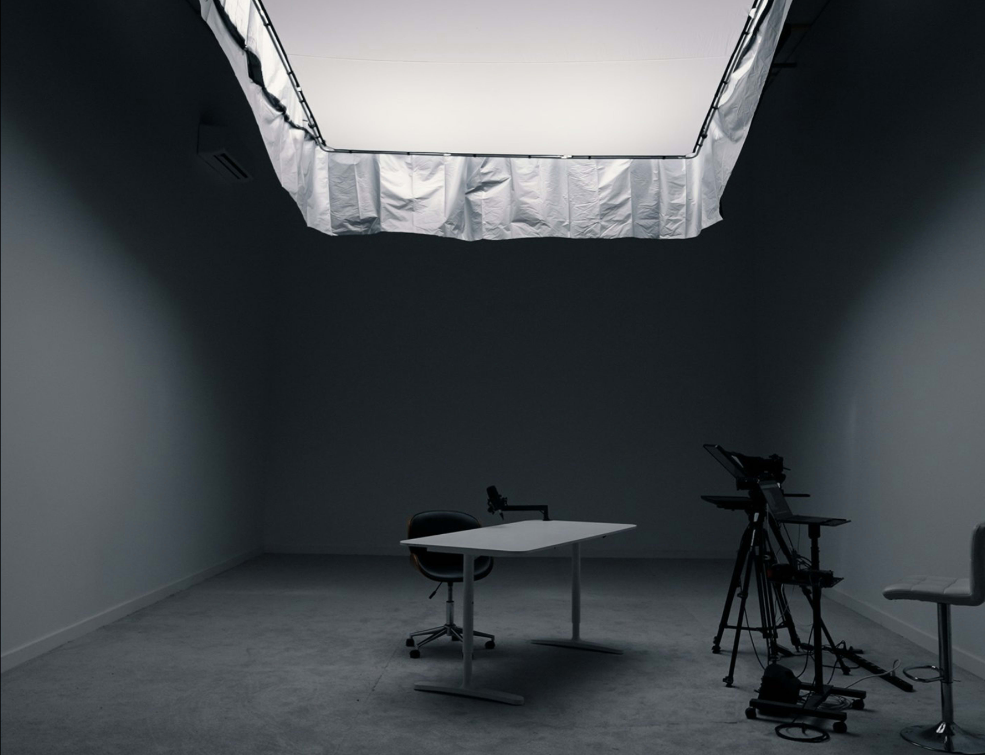 The image shows a dimly lit room with a single table and chair, alongside a camera setup in the corner.