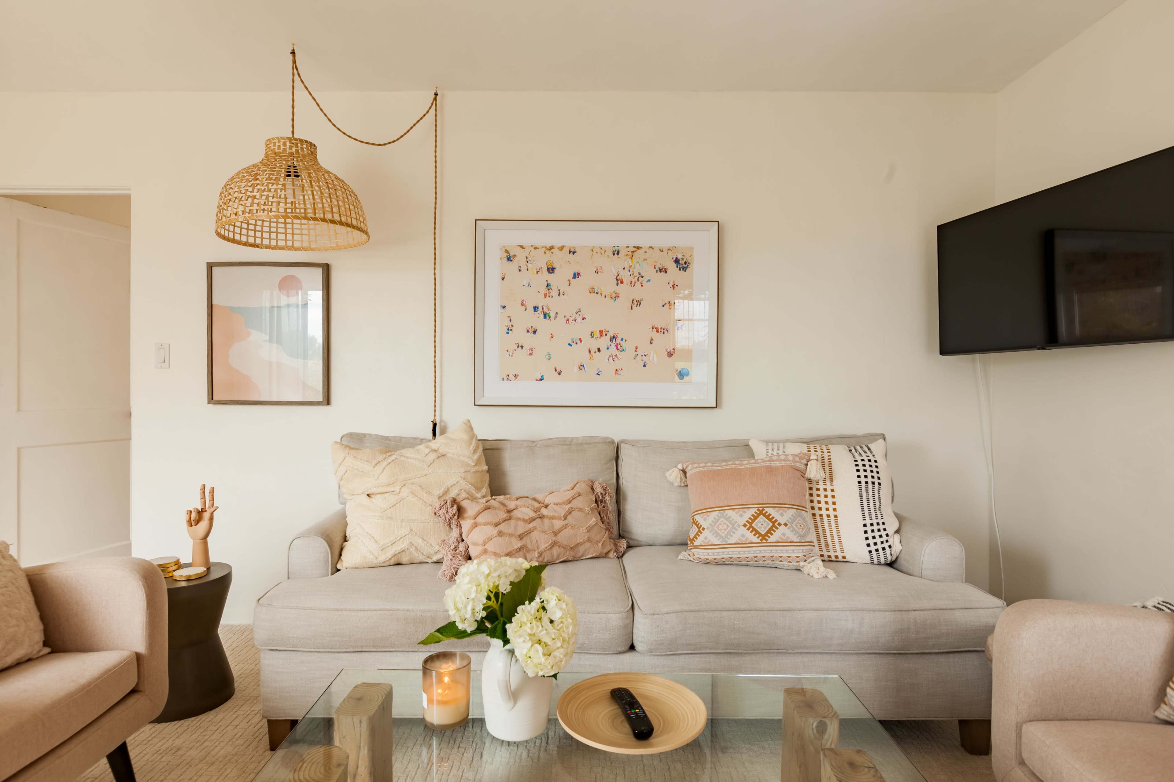 A cozy living room featuring a light-colored sofa, a glass coffee table with a candle and flowers, and a wall-mounted TV.