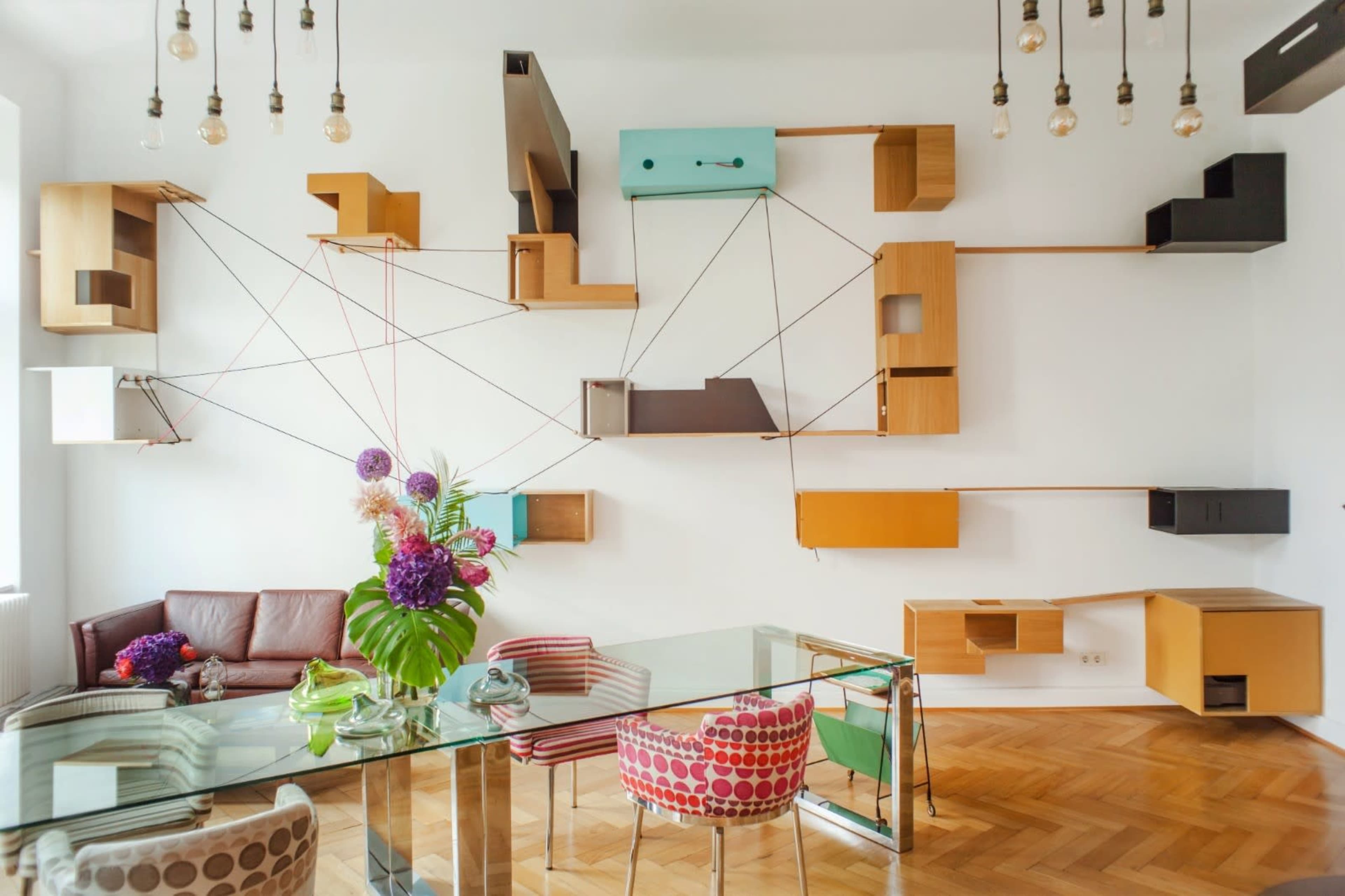 A modern living space with a glass dining table, colorful chairs, and an artistic wall display of wooden shelves connected by colorful cords.