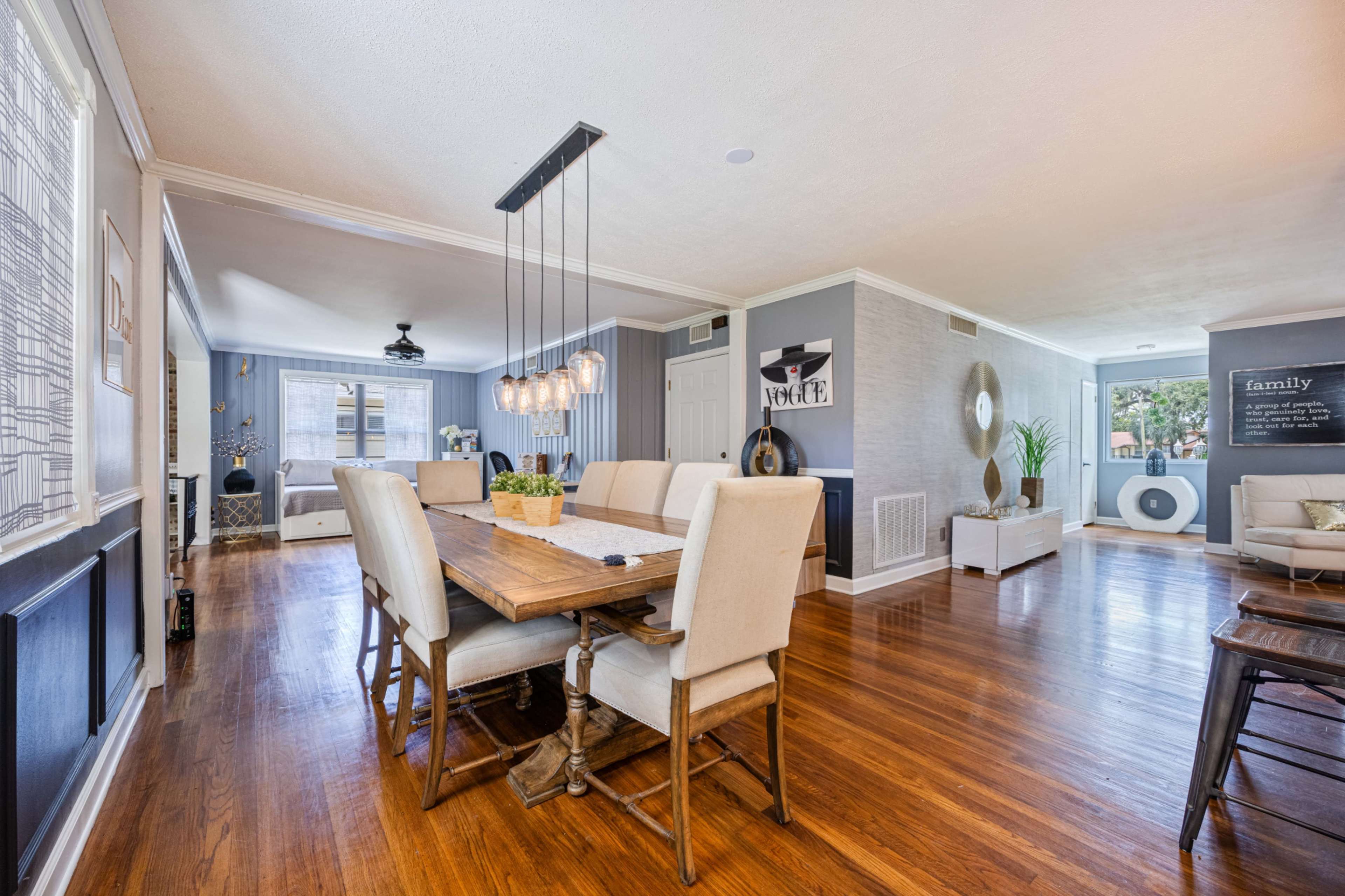 A modern dining area with a wooden table surrounded by upholstered chairs, illuminated by a pendant light, and leading into a living space.