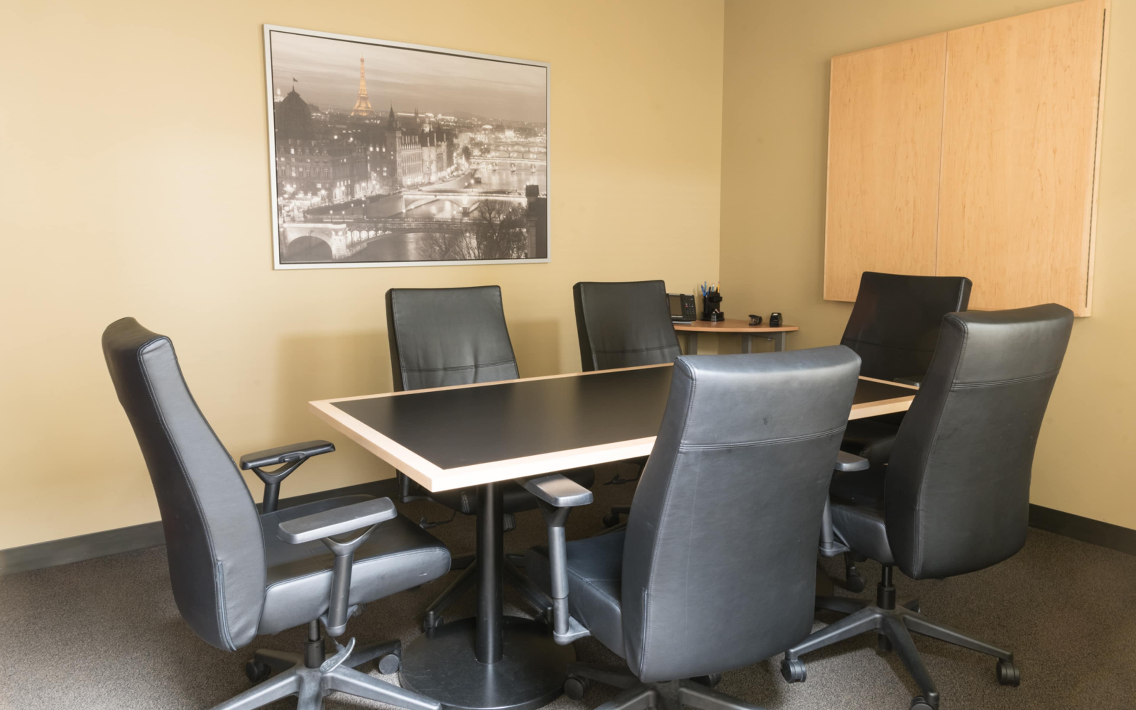 A conference room features a large rectangular table surrounded by six black leather chairs and a wall-mounted black-and-white photo of a cityscape.