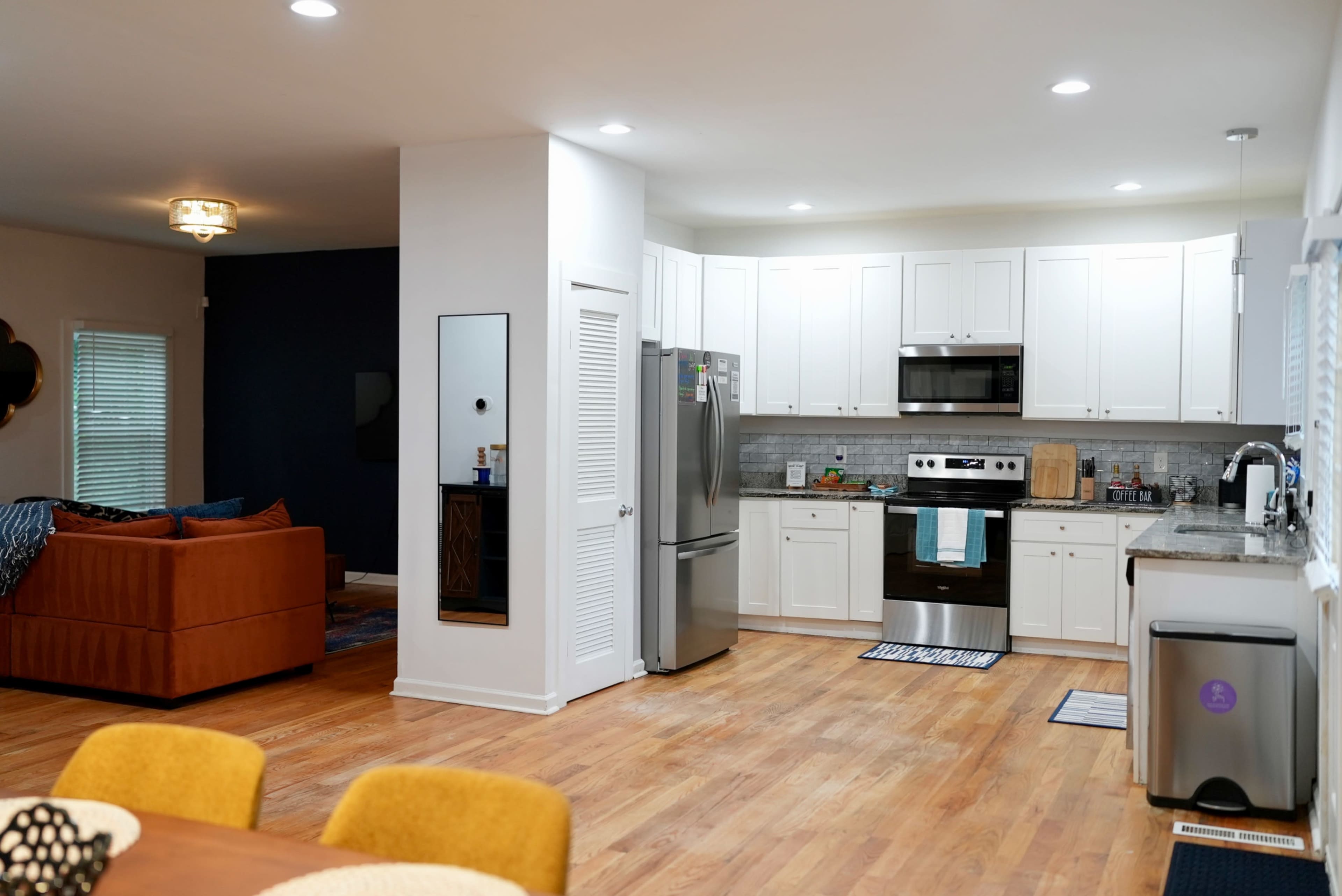 The image shows a modern kitchen with white cabinets, stainless steel appliances, and a dining area featuring a wooden table and colorful chairs.